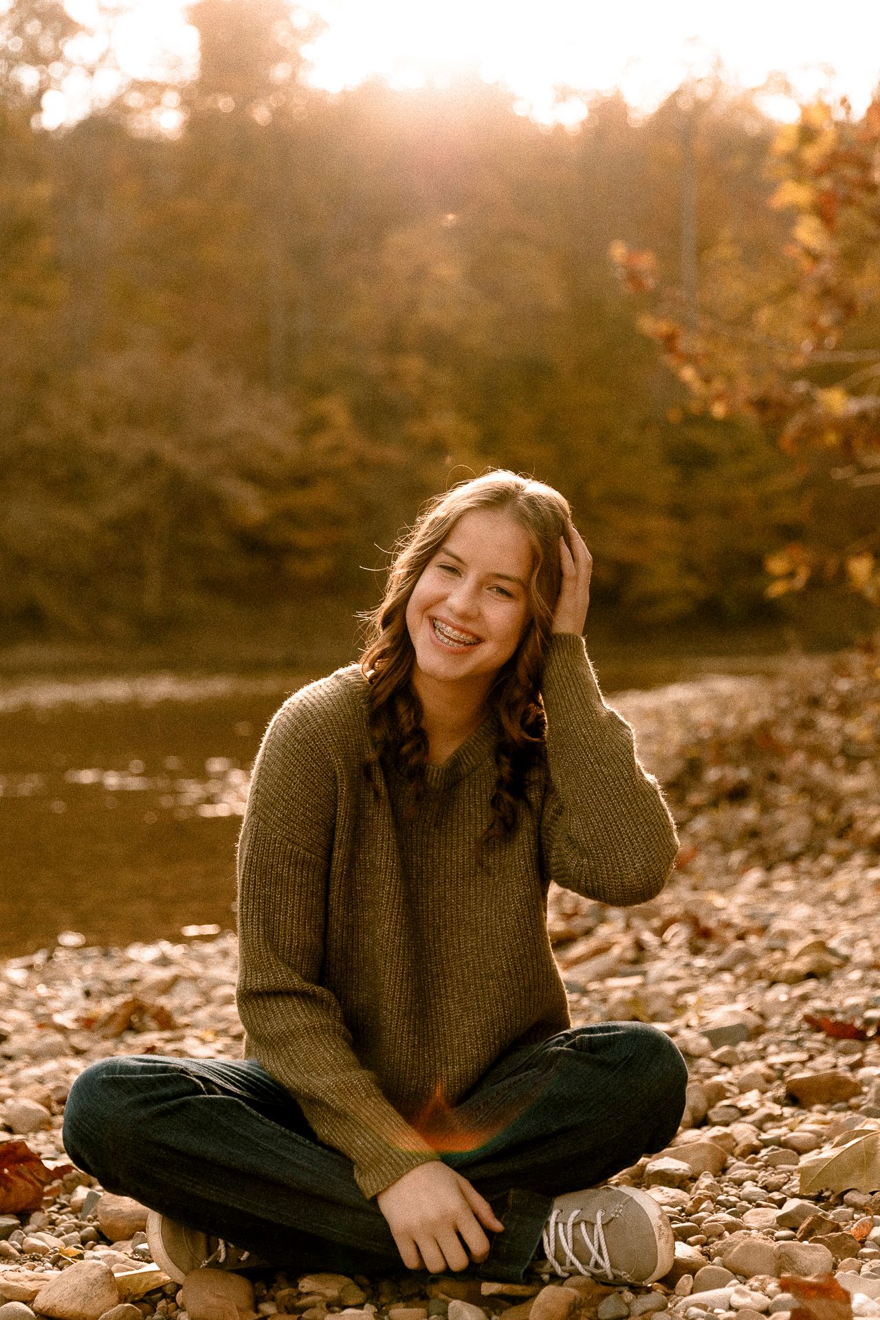 Young woman in olive sweater sitting cross-legged on rocky riverbank surrounded by autumn foliage in golden sunlight.
