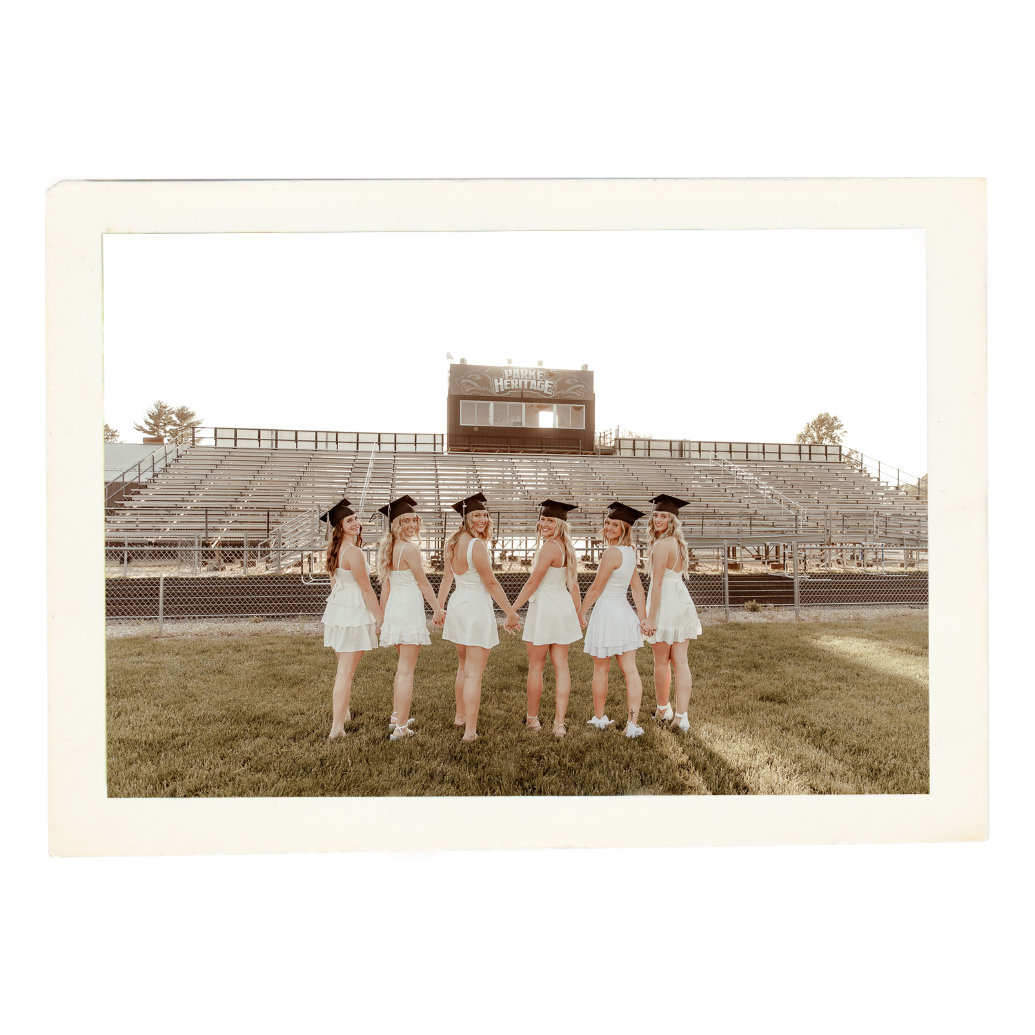 A group in matching white dresses stands in formation on a grassy field with stadium seating visible in the background.