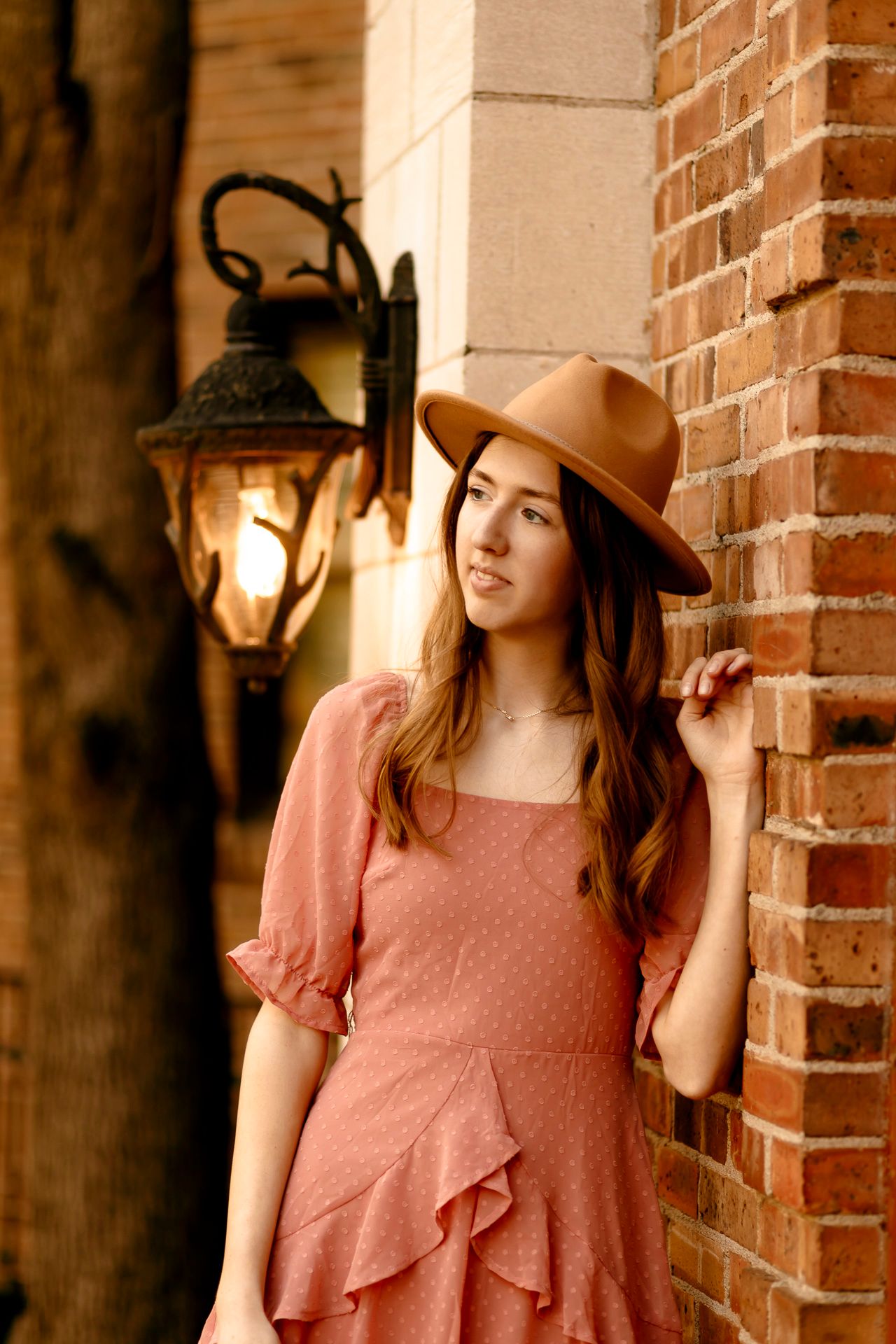 Young woman in pink ruffle dress and tan wide-brim hat leaning against brick wall near vintage lantern.