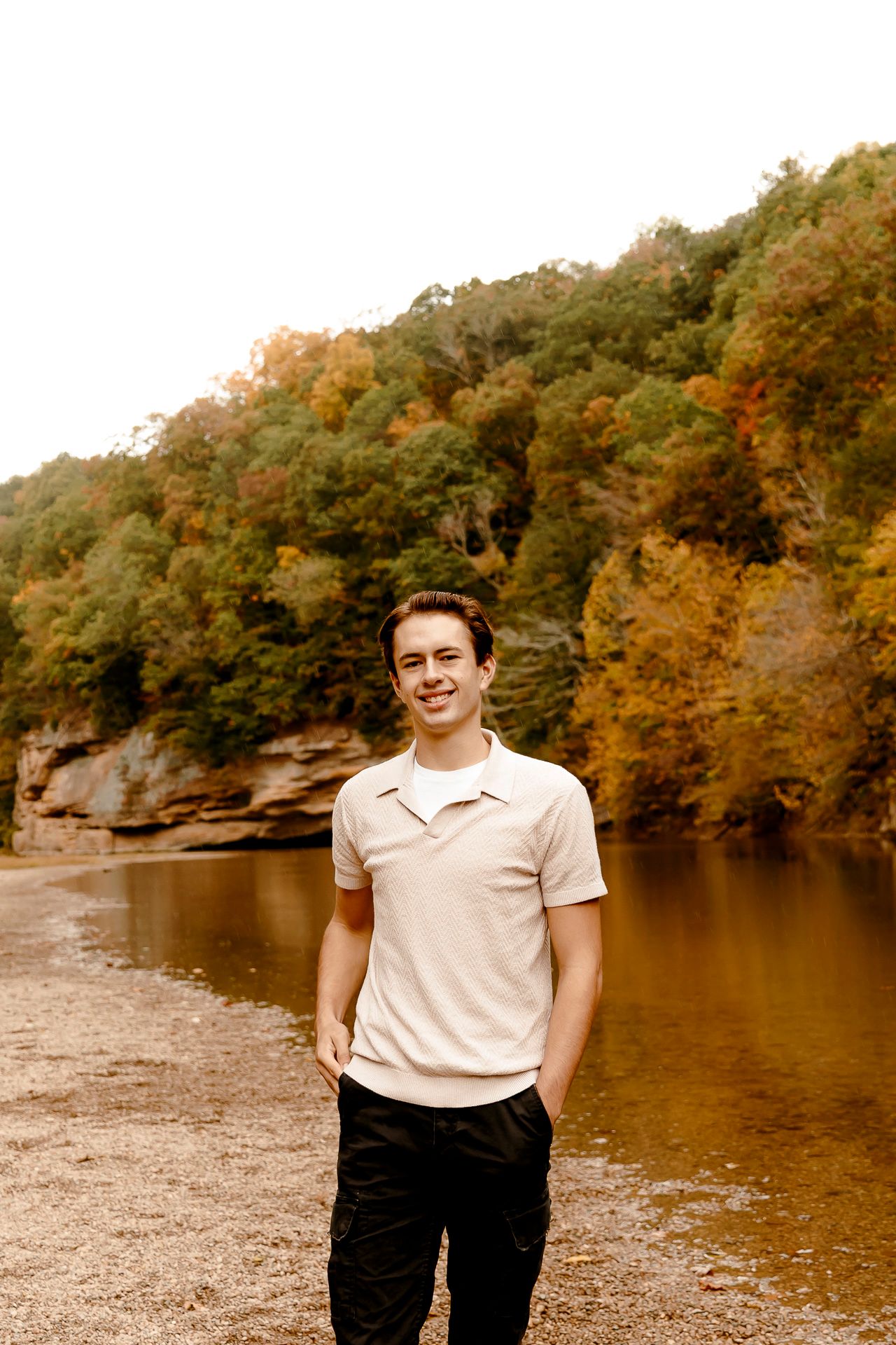Young man in beige polo smiles by a river with autumn foliage cliffs in background.