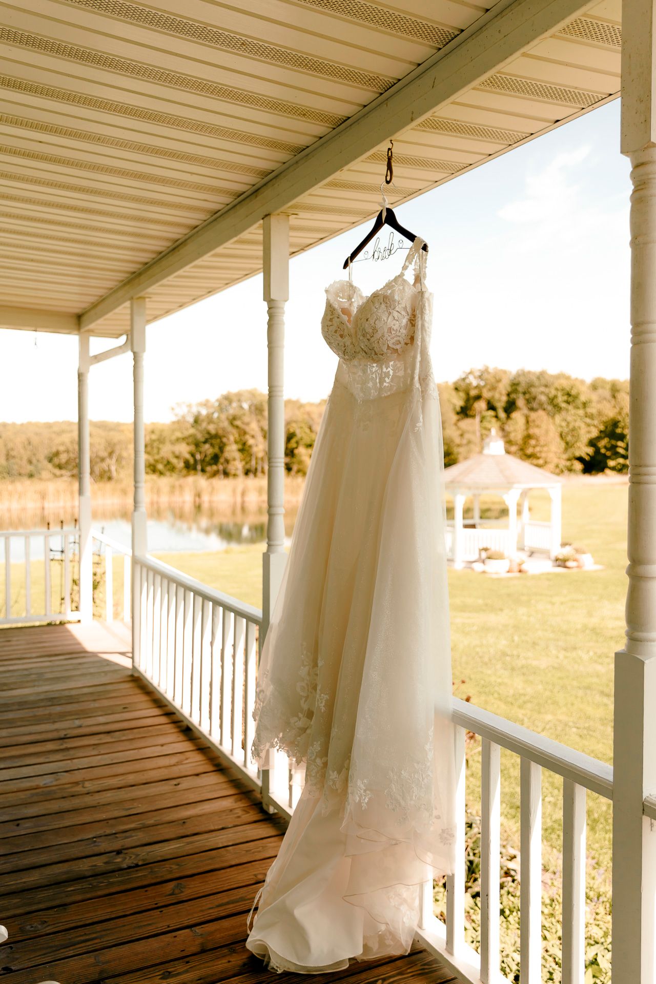 Elegant lace wedding dress hanging on a porch with a scenic gazebo and pond in the background.