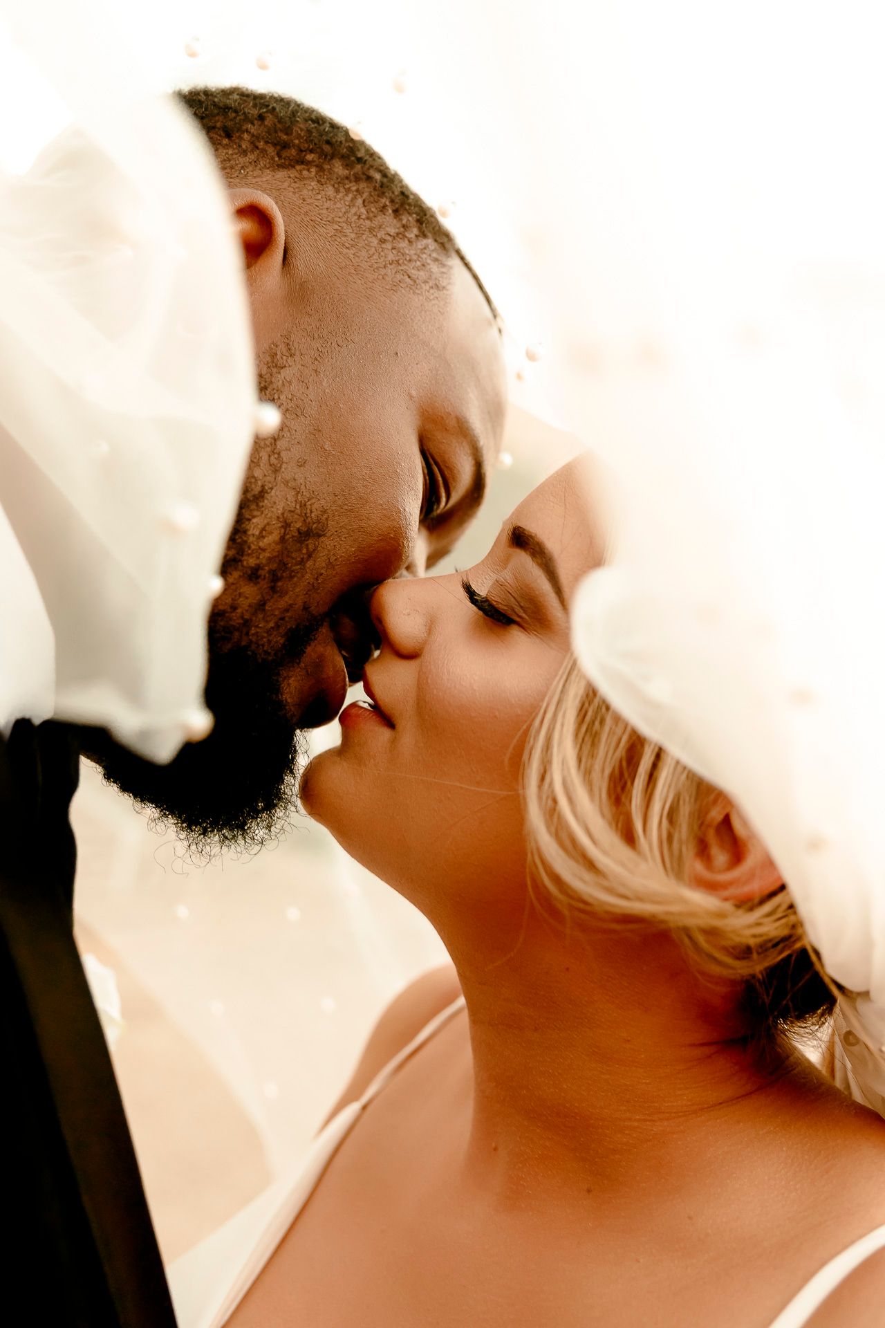 A romantic interracial wedding couple sharing a tender kiss under a flowing white bridal veil.