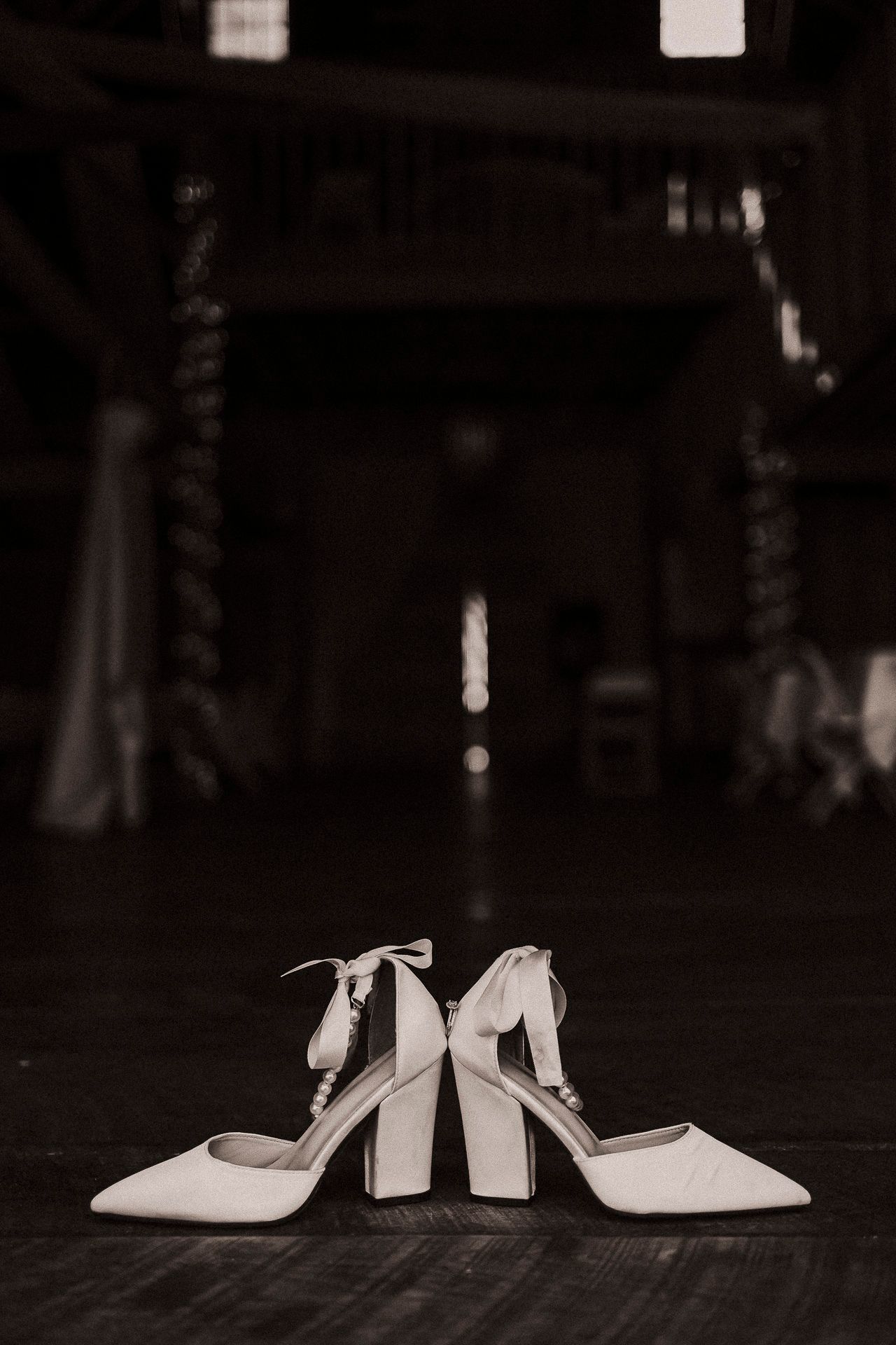 Elegant white bridal heels with ankle ties displayed on dark floor in black and white wedding photography.