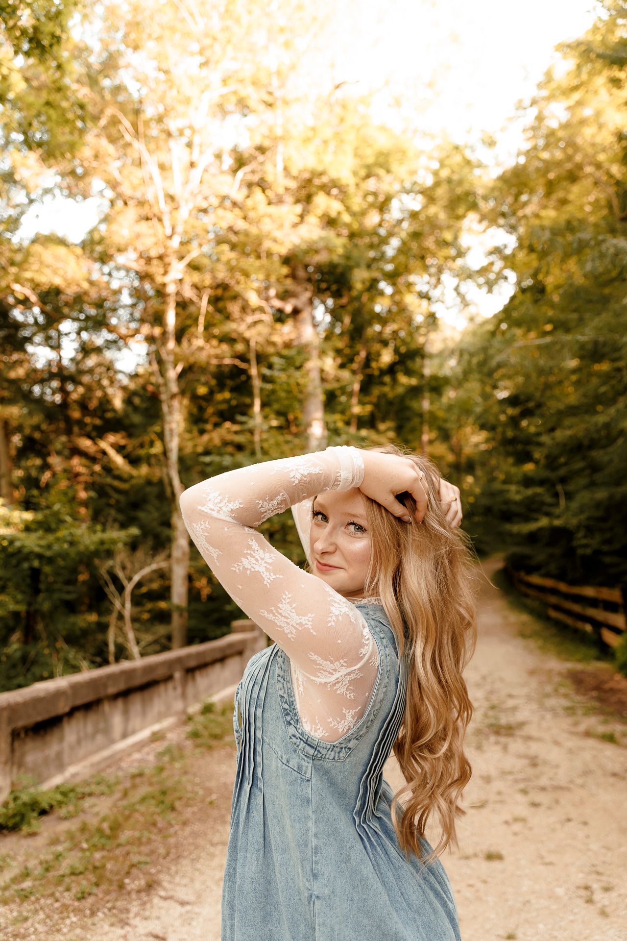 Young woman in blue dress with lace sleeves poses on a sunlit forest path, looking back over her shoulder.