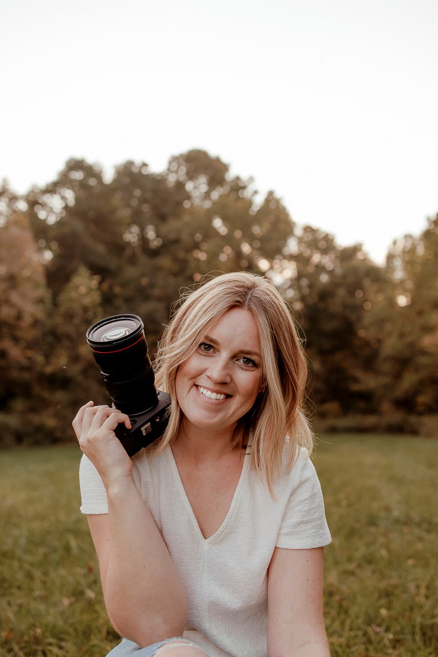 A photographer in a white shirt holds a camera while sitting in a grassy field at sunset.