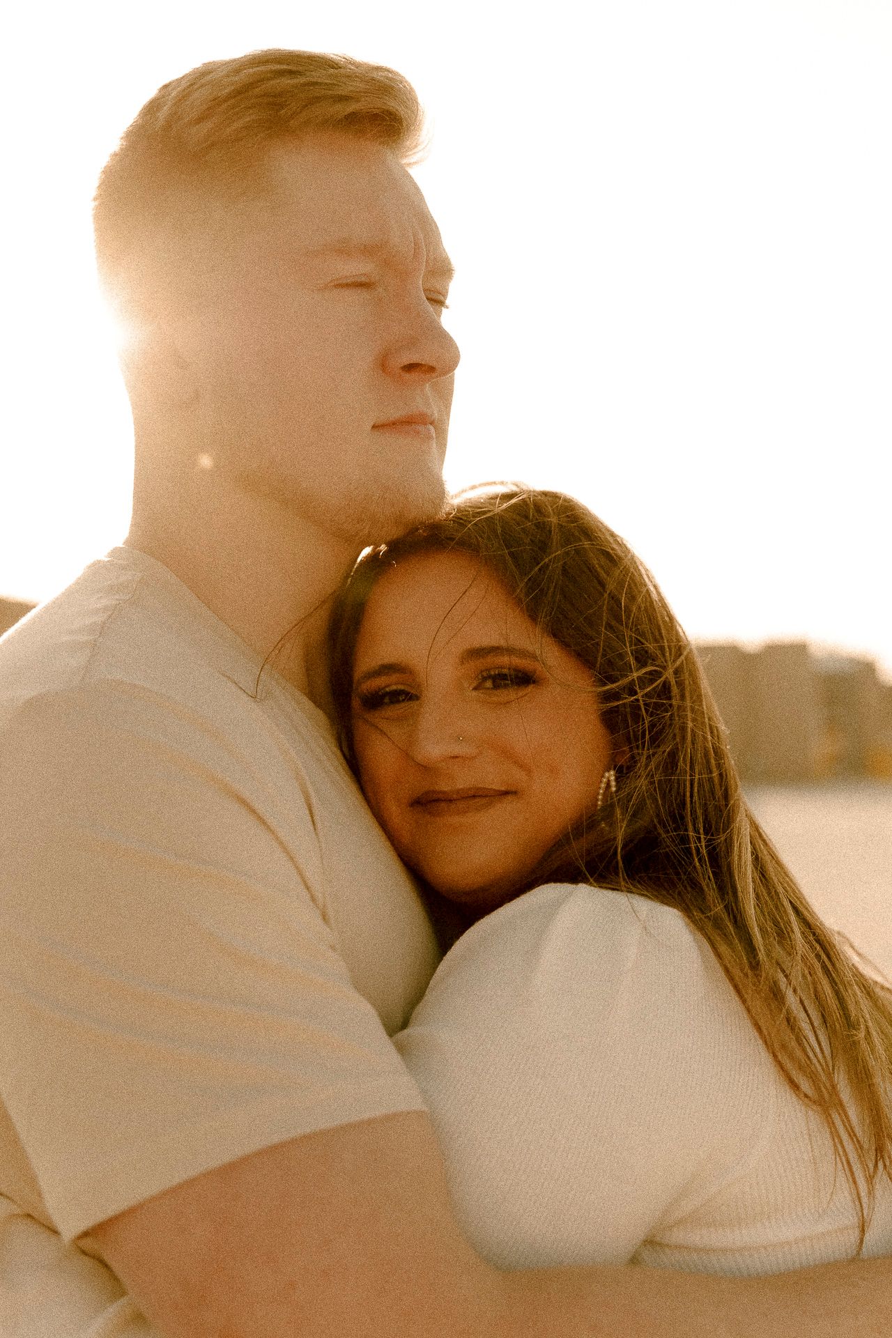 A couple embraces warmly at golden hour, woman smiling softly at camera while man holds her with eyes closed.