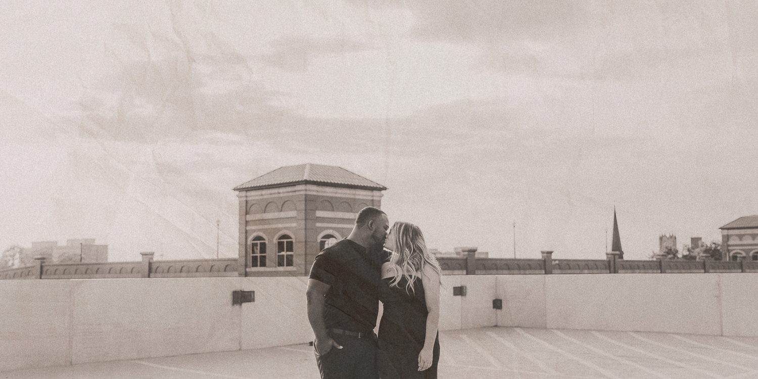 A couple stands together on a rooftop overlooking a city skyline in a vintage-style black and white photograph.