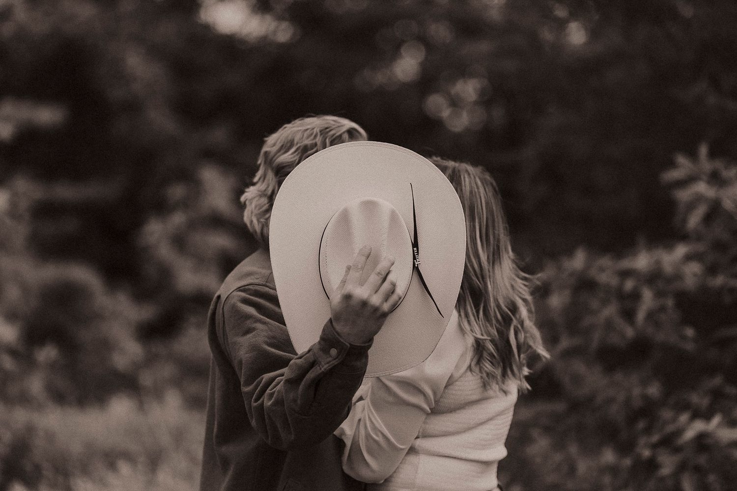 Couple sharing a romantic kiss hidden behind a wide-brim hat outdoors in a black and white photo.