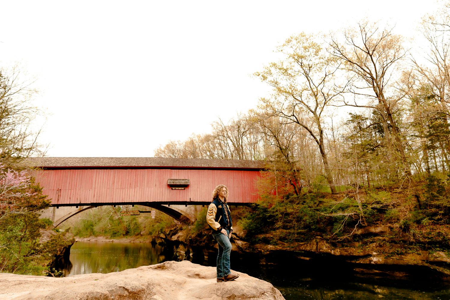 Person standing on rocks beside a red covered bridge over a calm river surrounded by autumn trees.