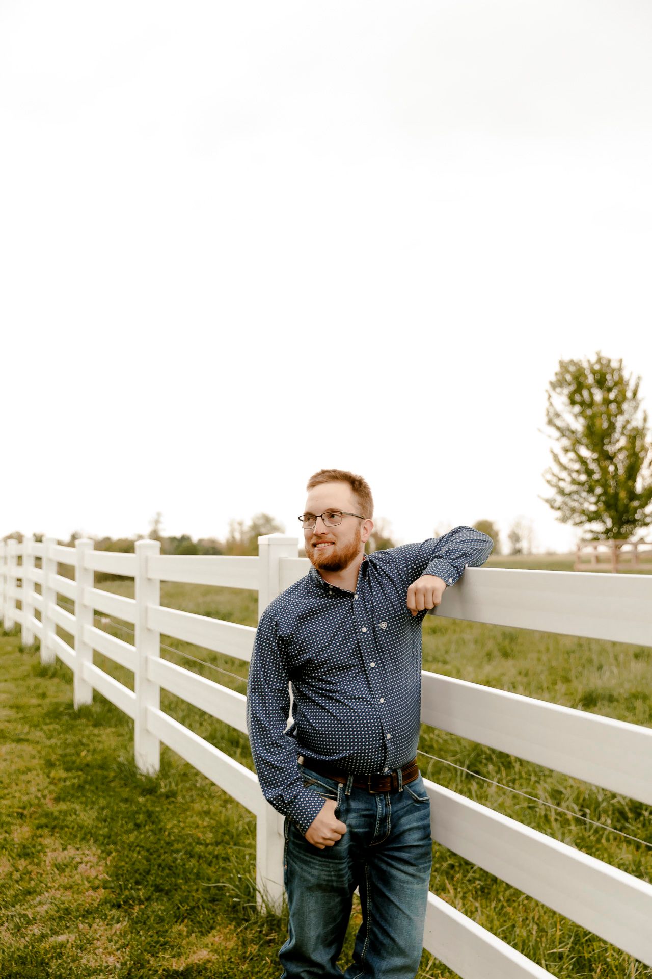 A bearded man in glasses and blue shirt leans against a white farm fence in a green rural landscape.