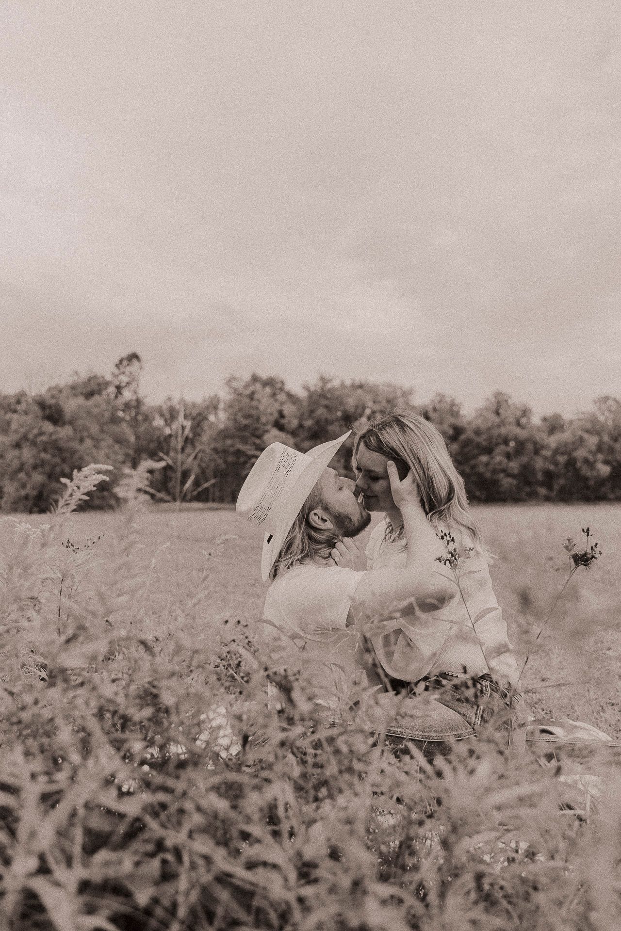 A black and white photograph showing two people embracing in a wildflower field on an overcast day.