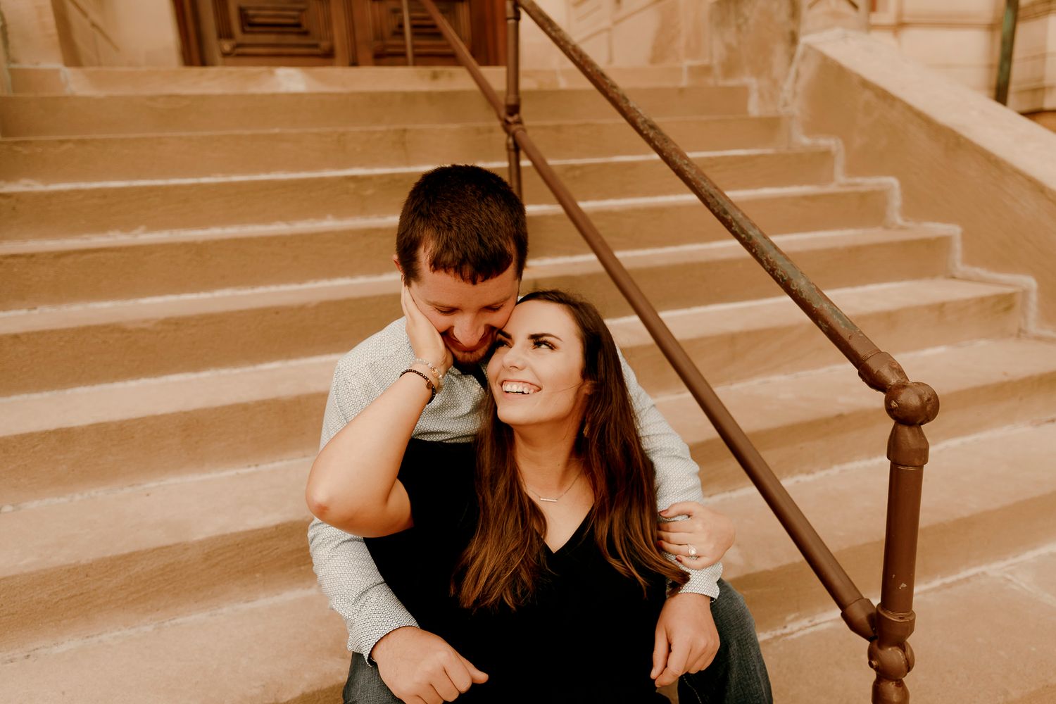 Couple sitting on stone steps, man whispering to smiling woman in black top during romantic engagement photo session.