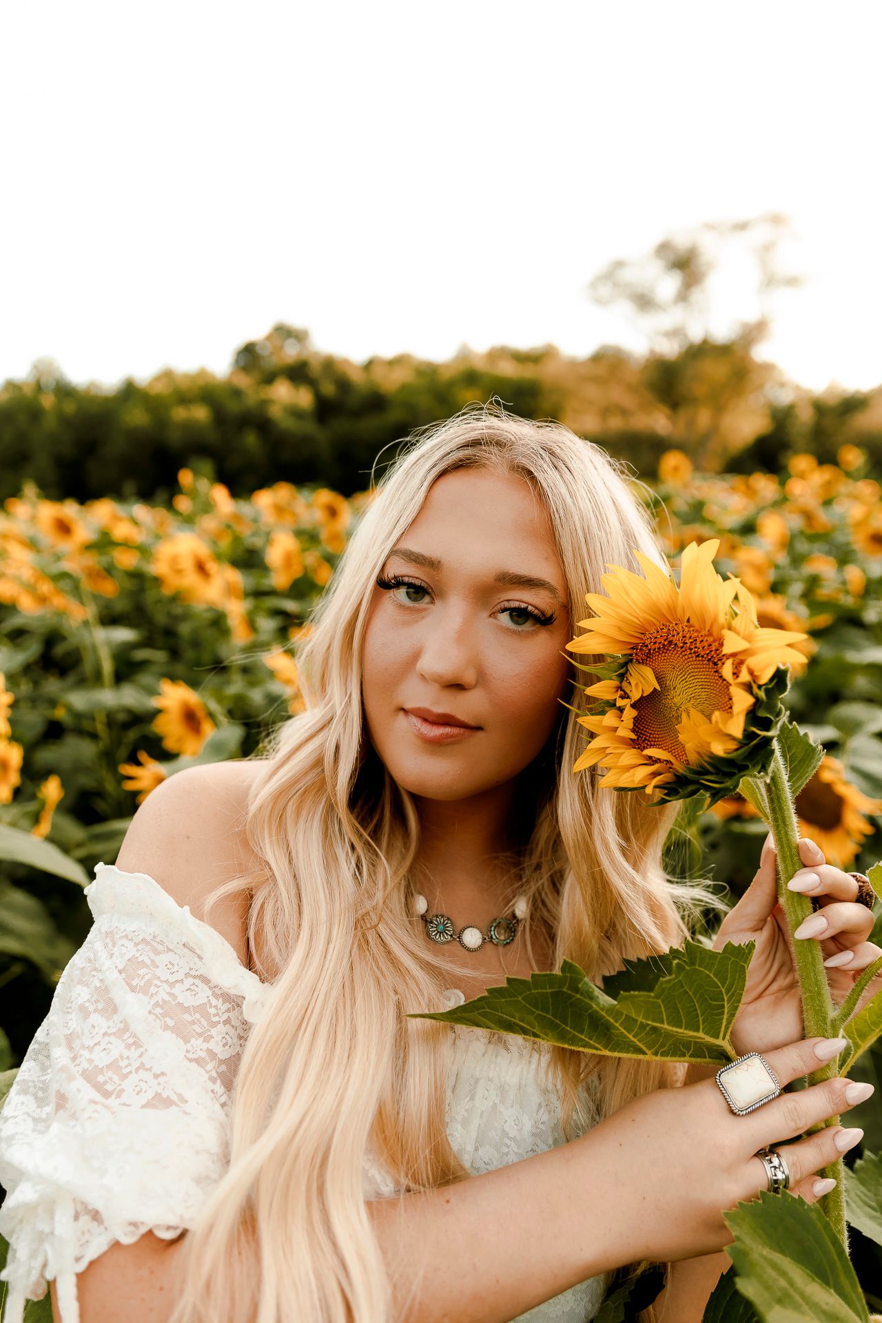 Young blonde woman holding a sunflower in a vibrant sunflower field, wearing a white lace dress and silver jewelry.
