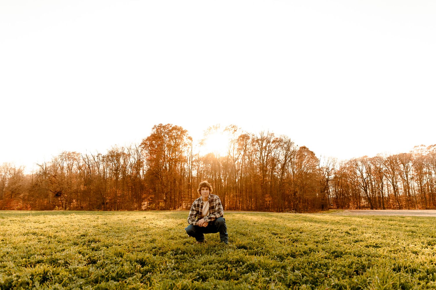 Person crouching in a sunlit green field with autumn trees glowing in golden hour light in the background.