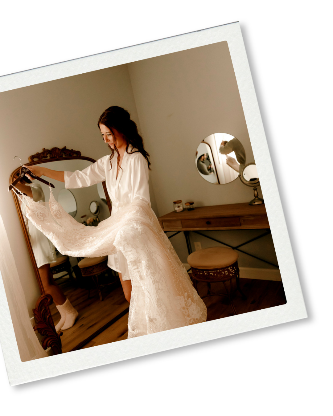 Bride in white lace robe holding wedding dress in front of ornate mirror during bridal preparation.