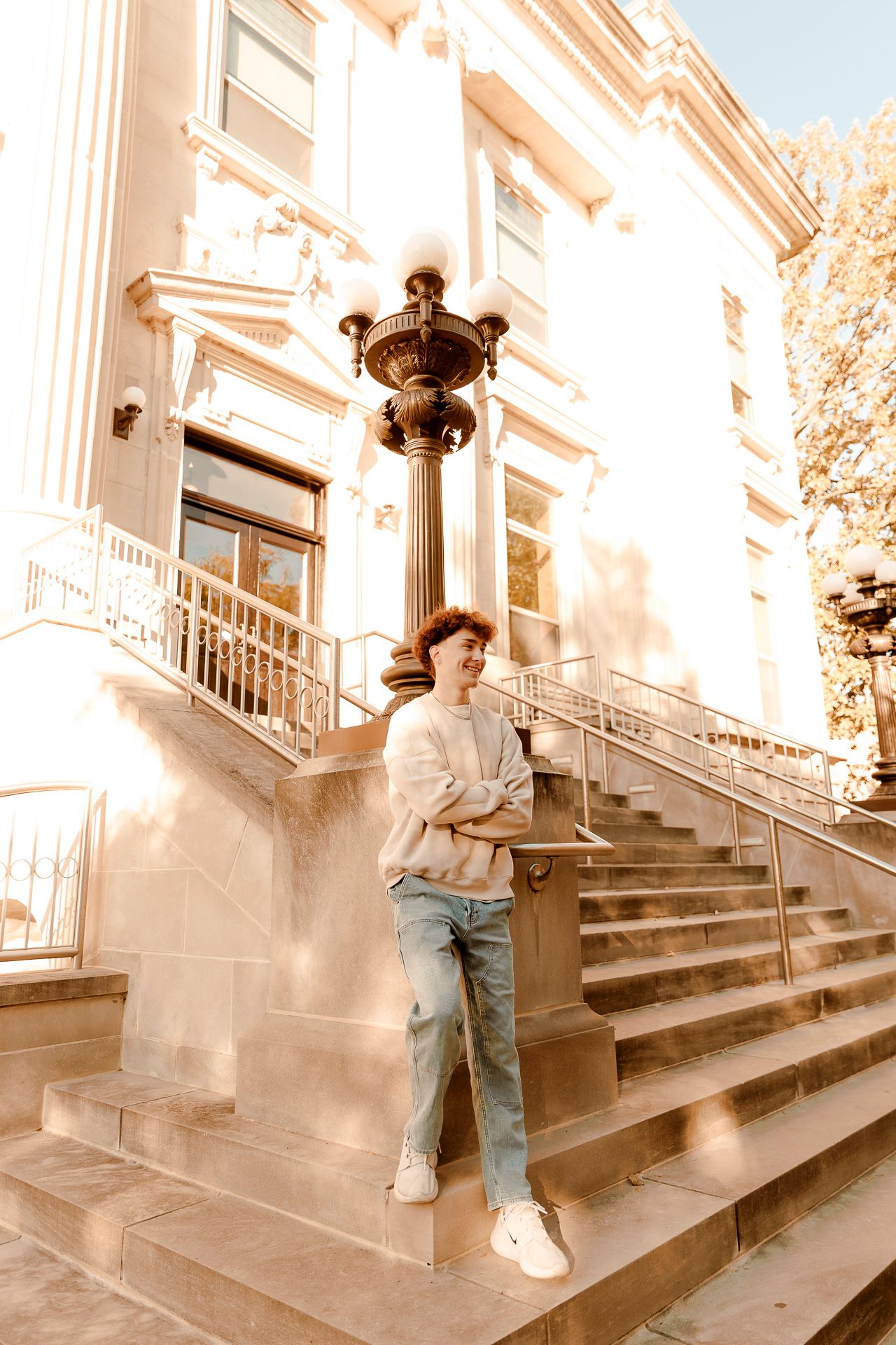 Young man in jeans and sweatshirt leaning against ornate street lamp on steps of classic building.