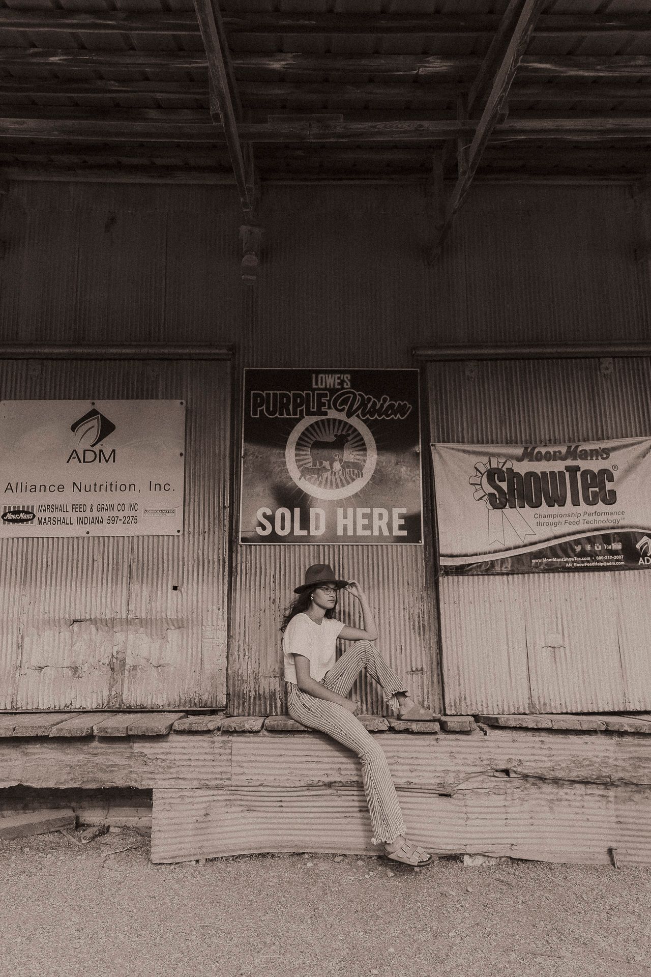Woman in cowboy hat sits on ledge before rustic barn with vintage ADM and ShowTec signs in sepia tone.