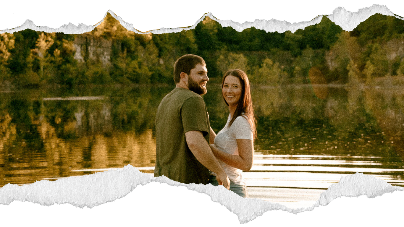 A couple smiles and embraces on a dock by a reflective lake surrounded by autumn trees and rocky cliffs.