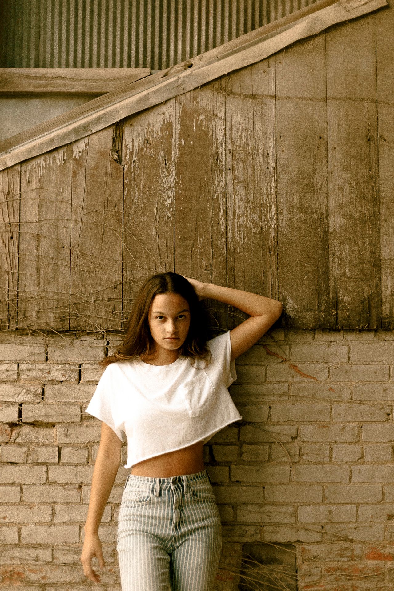 Young woman in white crop top and striped jeans leaning against rustic brick wall with weathered wood background.