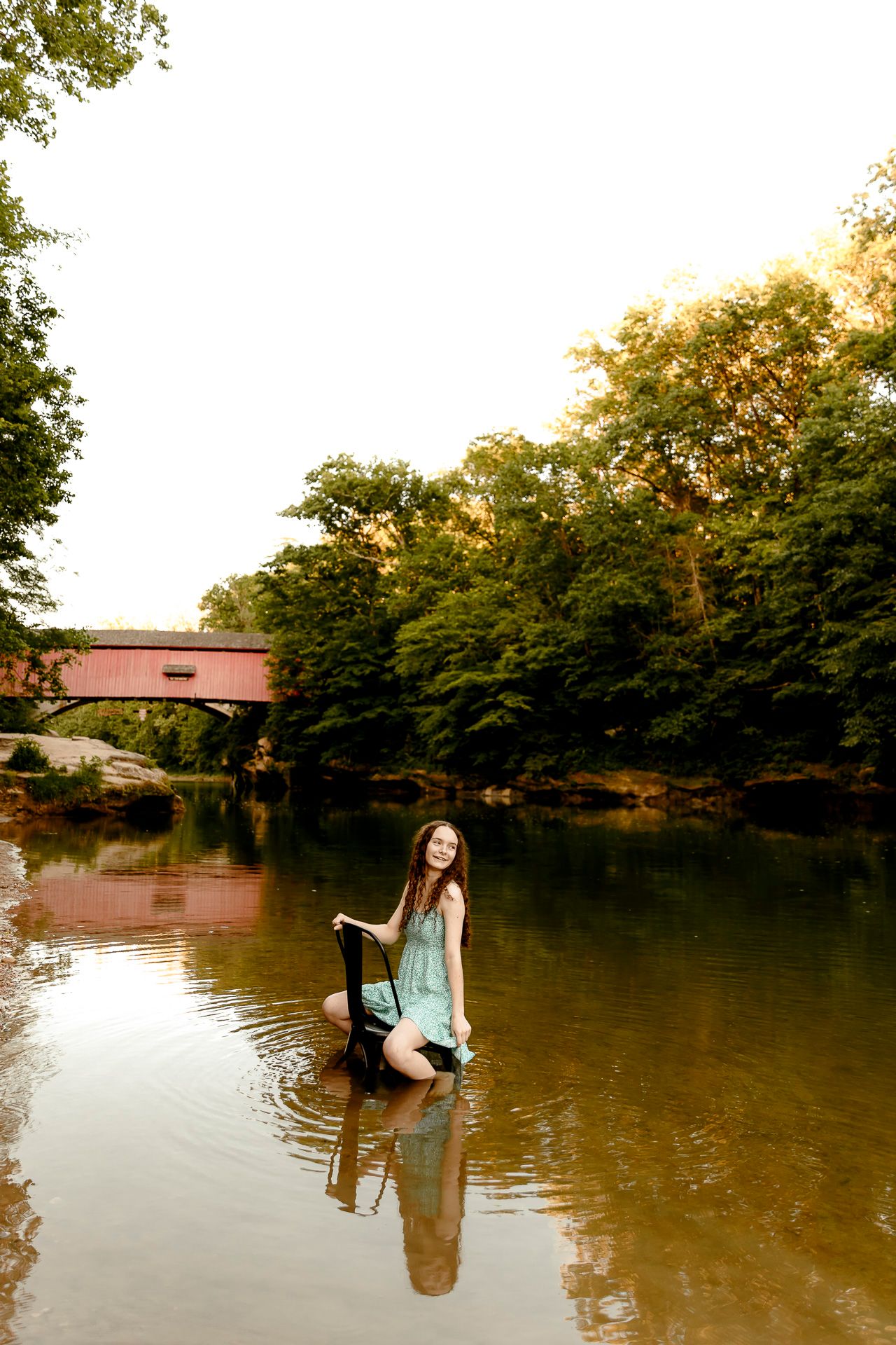A young woman in a teal dress stands in a shallow river holding a book, with a red covered bridge visible among trees.