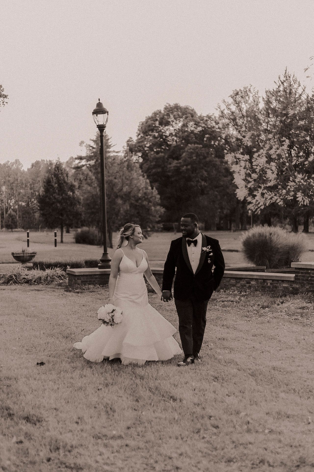 Bride and groom walk hand-in-hand through a lush park in black and white wedding photo.