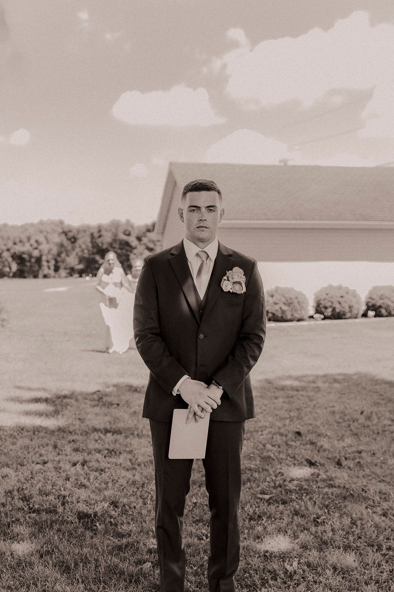 Groom in dark suit holding vows card outdoors, bride in white dress visible in background, sepia-toned wedding photo.