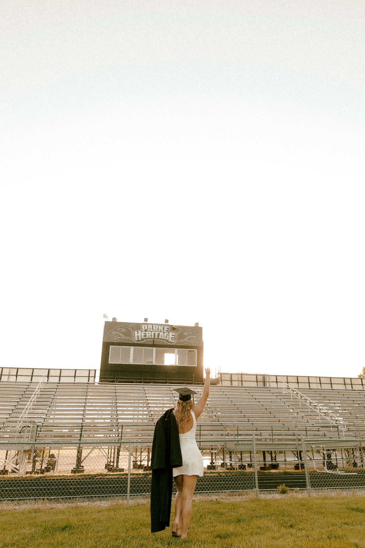 Graduate in white dress holds cap up at Hawks Heritage stadium bleachers during golden hour celebration.