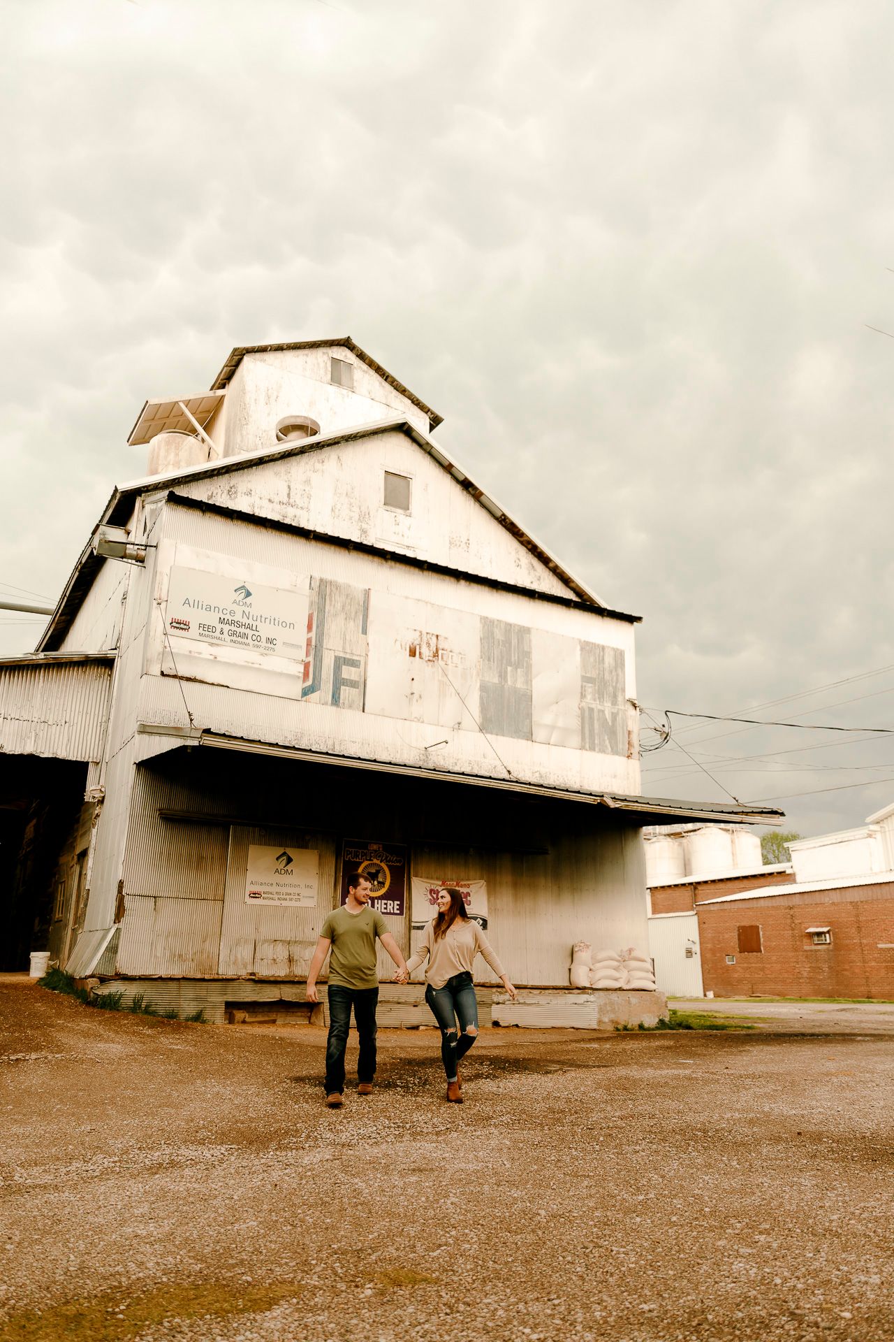 Couple holding hands walking in front of a rustic white grain elevator building under dramatic cloudy skies.