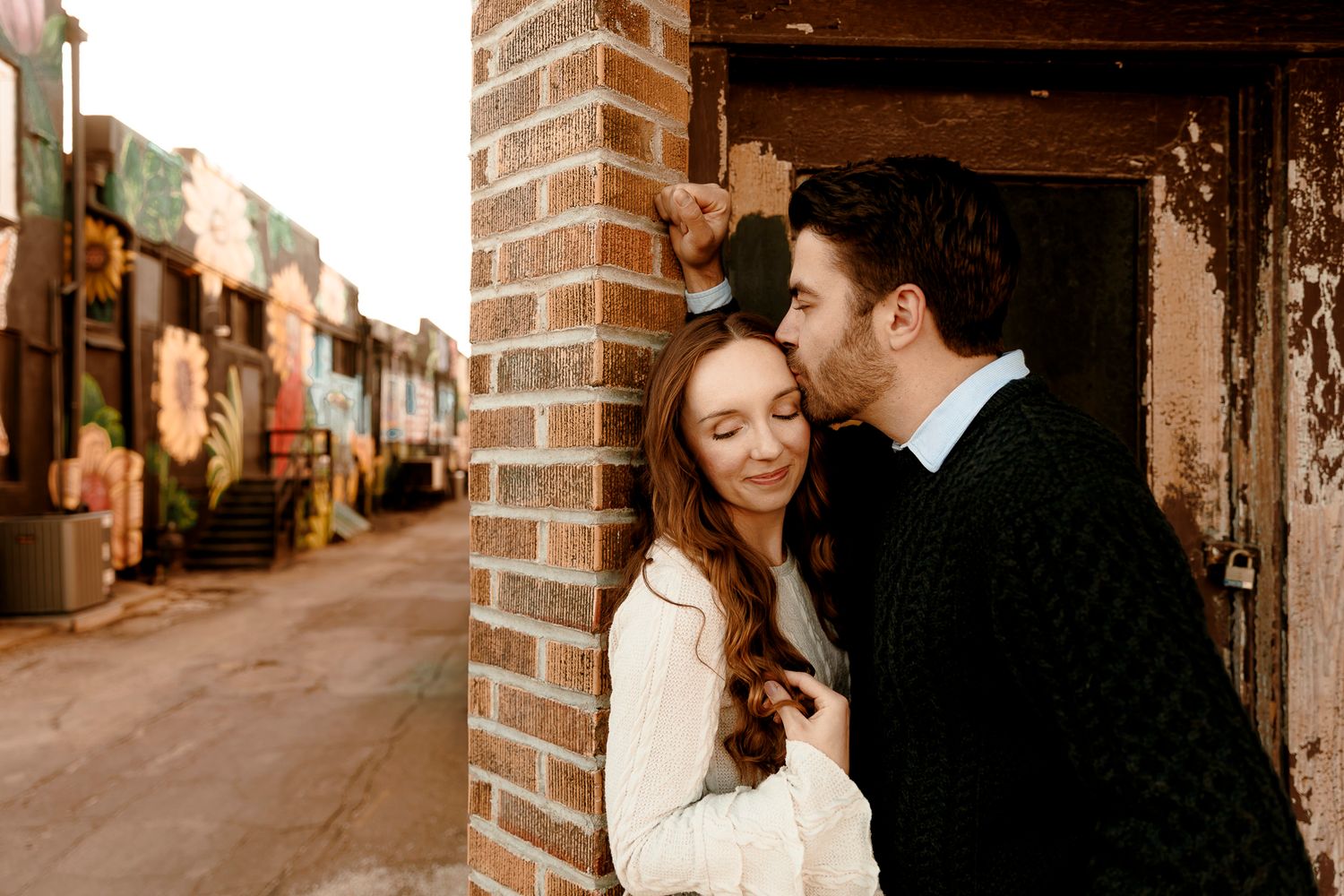Couple sharing a romantic moment by a brick pillar in an urban alley with colorful graffiti murals in the background.