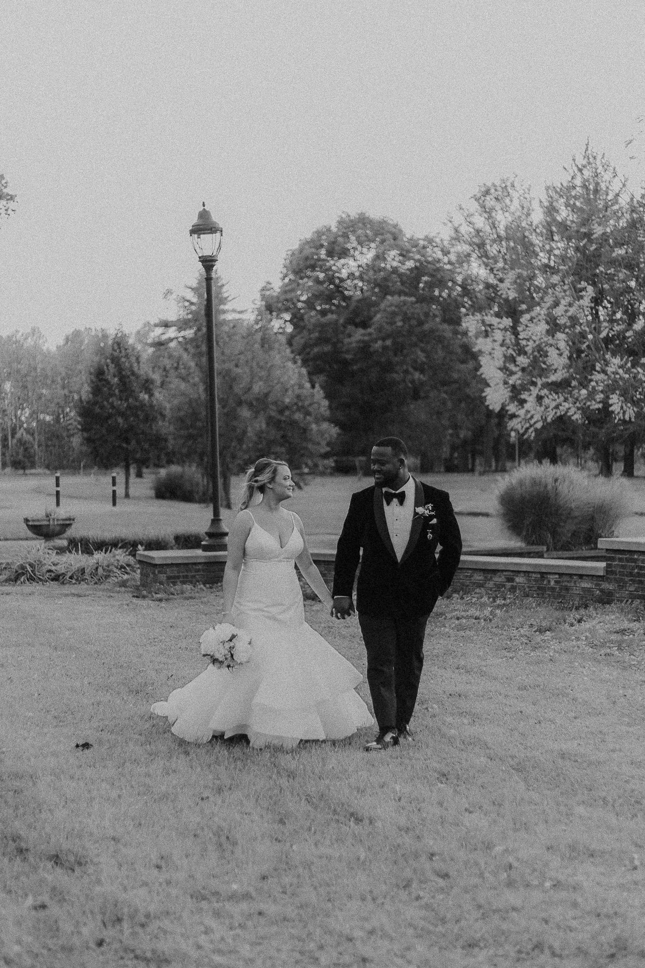 A couple in formal wedding attire walking together across a grassy field in black and white photography.