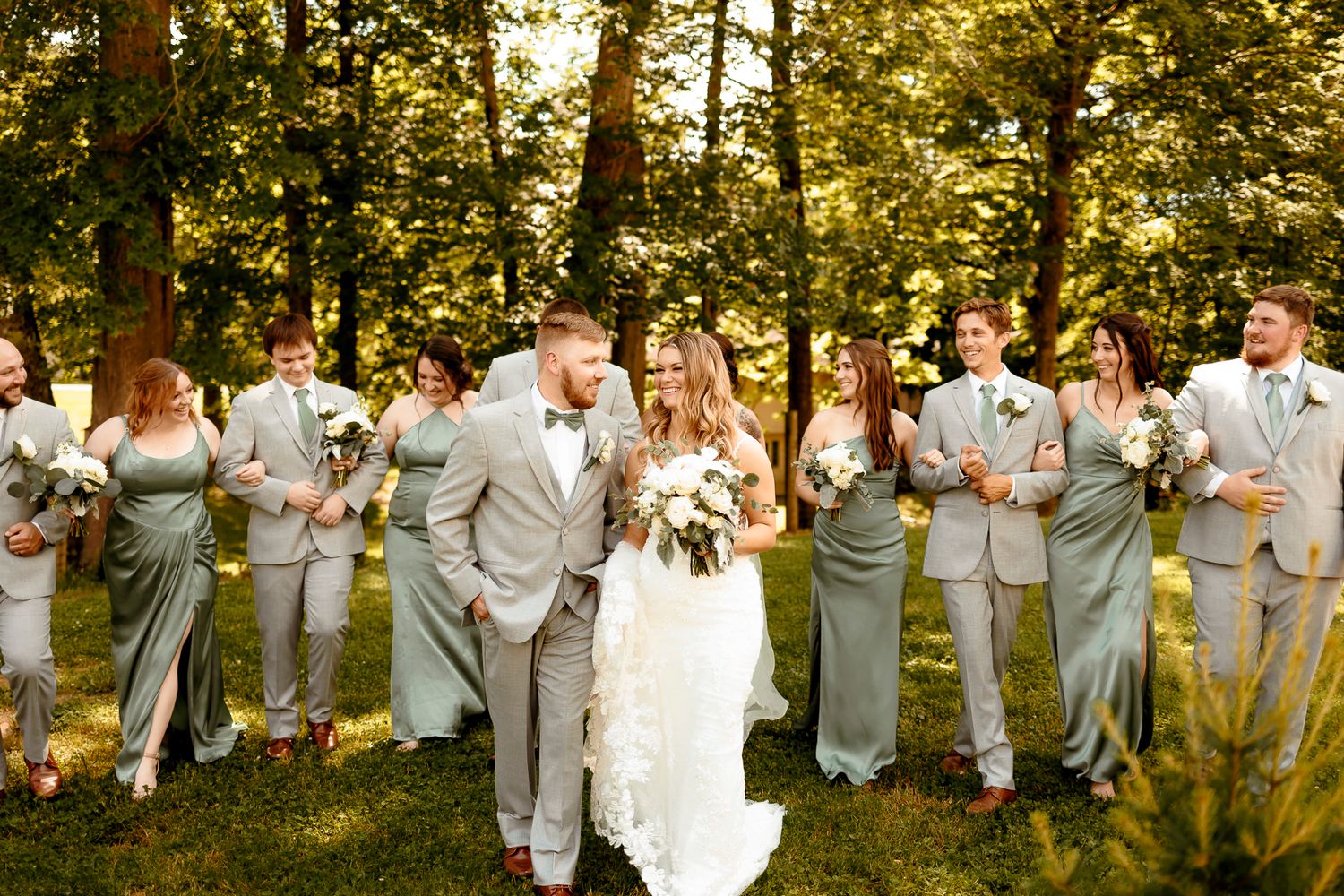 Wedding party in sage green attire poses outdoors among lush trees, bride and groom sharing a romantic moment.