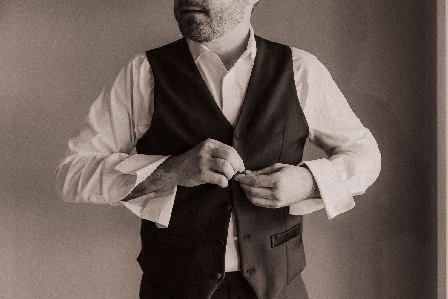Black and white photo of a groom buttoning his vest while getting ready for his wedding ceremony.