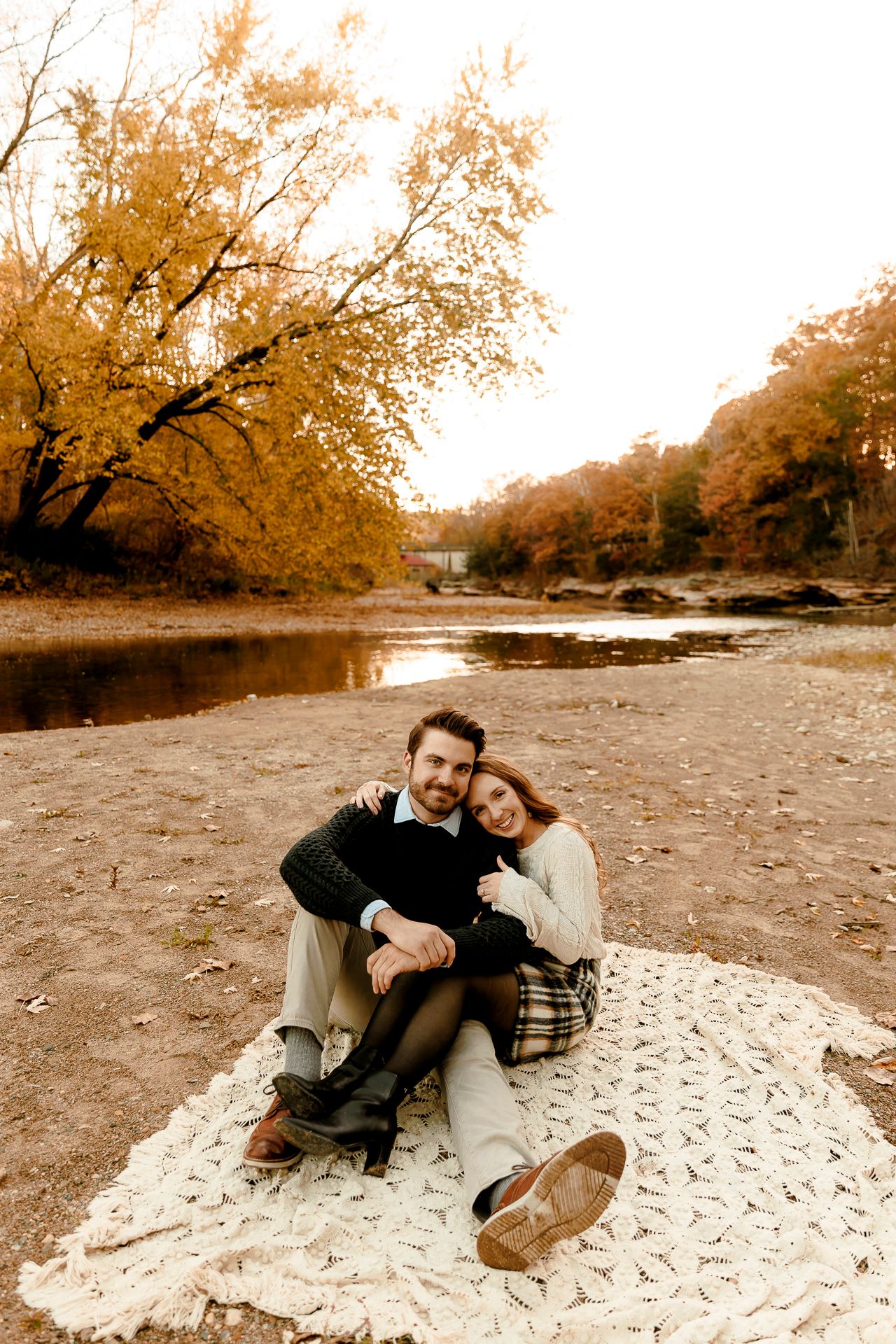 Couple sitting on blanket by autumn river, surrounded by golden fall foliage, sharing a romantic embrace outdoors.