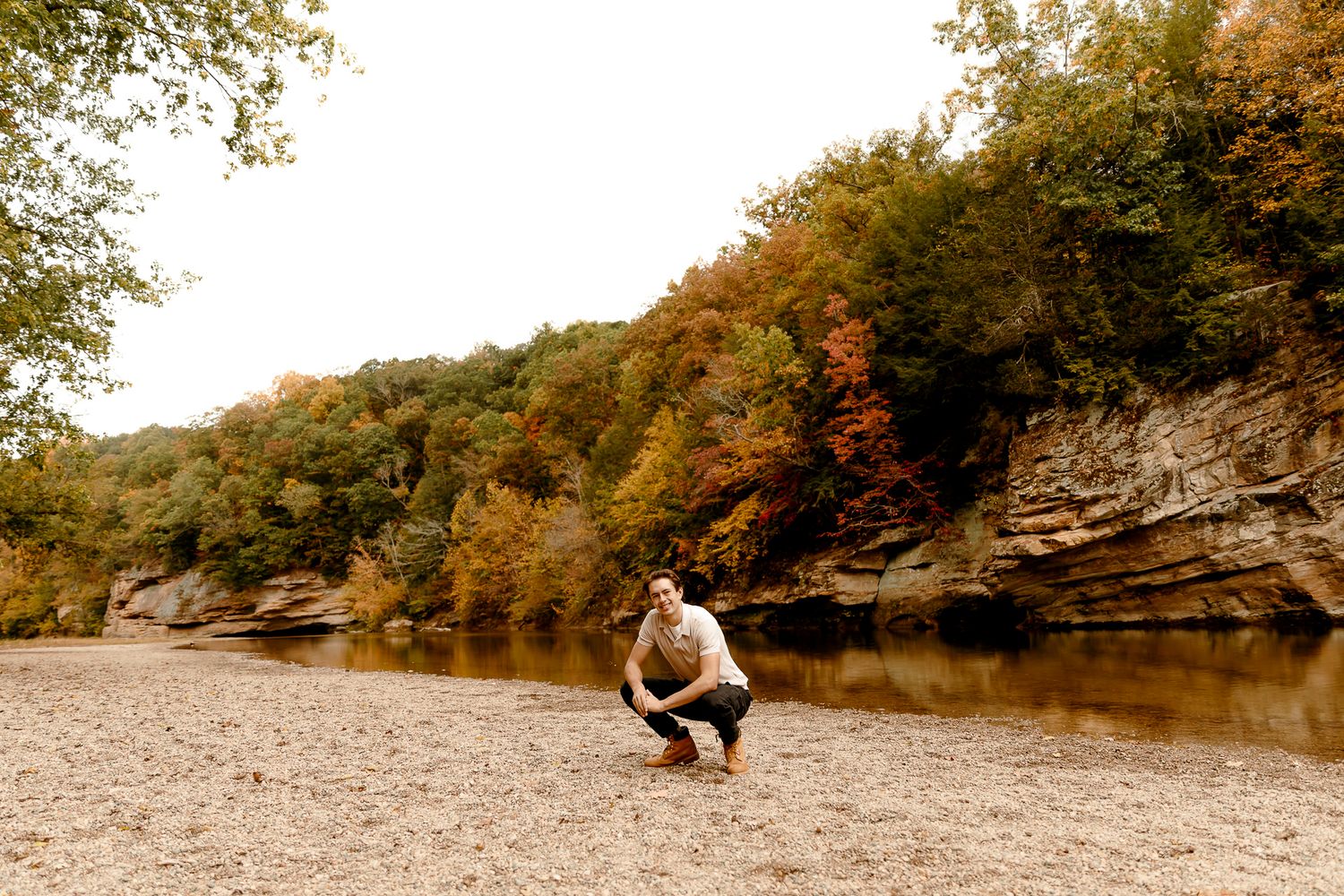 Man crouching on rocky riverbank surrounded by autumn foliage and sandstone cliffs in fall landscape.
