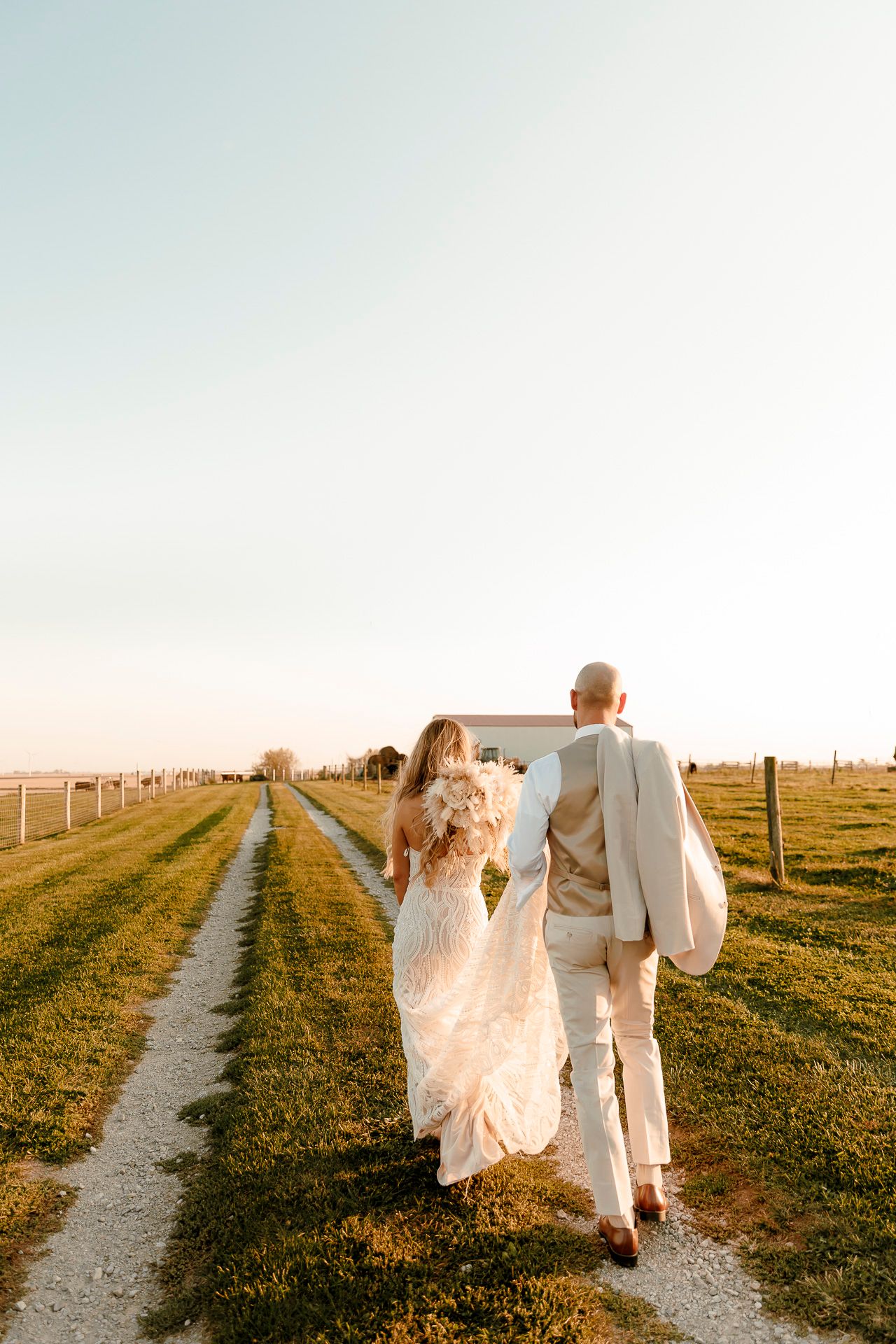 Bride and groom walking hand in hand down a rural path at golden hour, surrounded by green fields.
