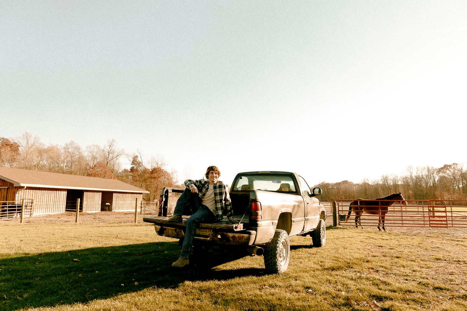 Person sitting on pickup truck tailgate at a rural horse farm with stables and horses in background.
