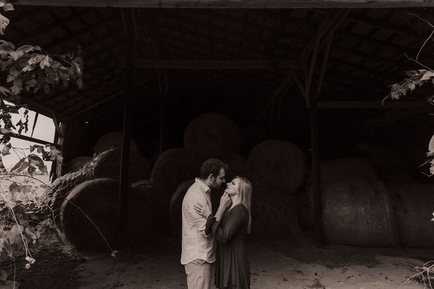 Couple sharing a romantic moment in front of a rustic barn filled with hay bales in a black and white photo.