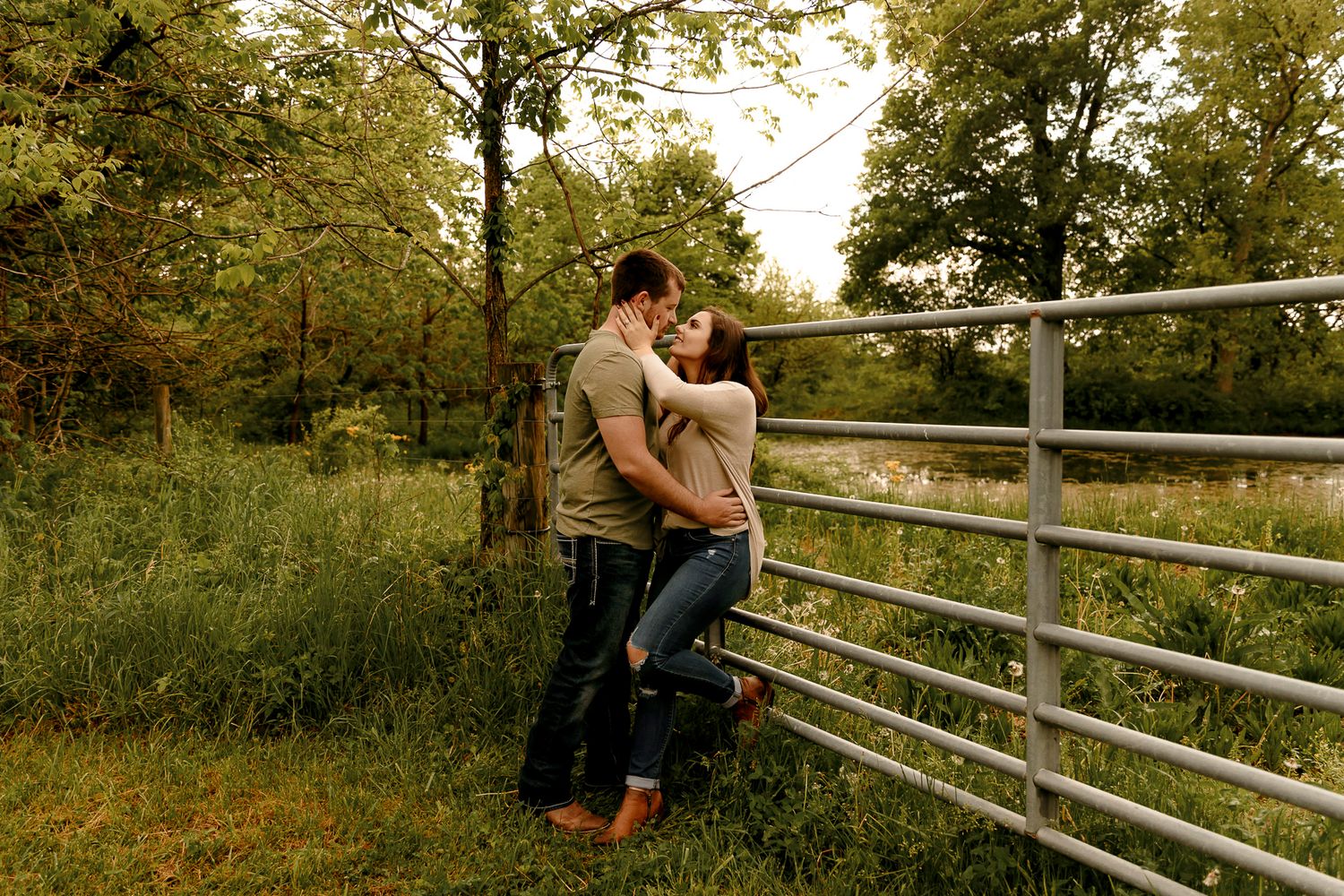 Couple embracing romantically by a rustic metal fence surrounded by lush green trees and tall grass in a countryside setting.