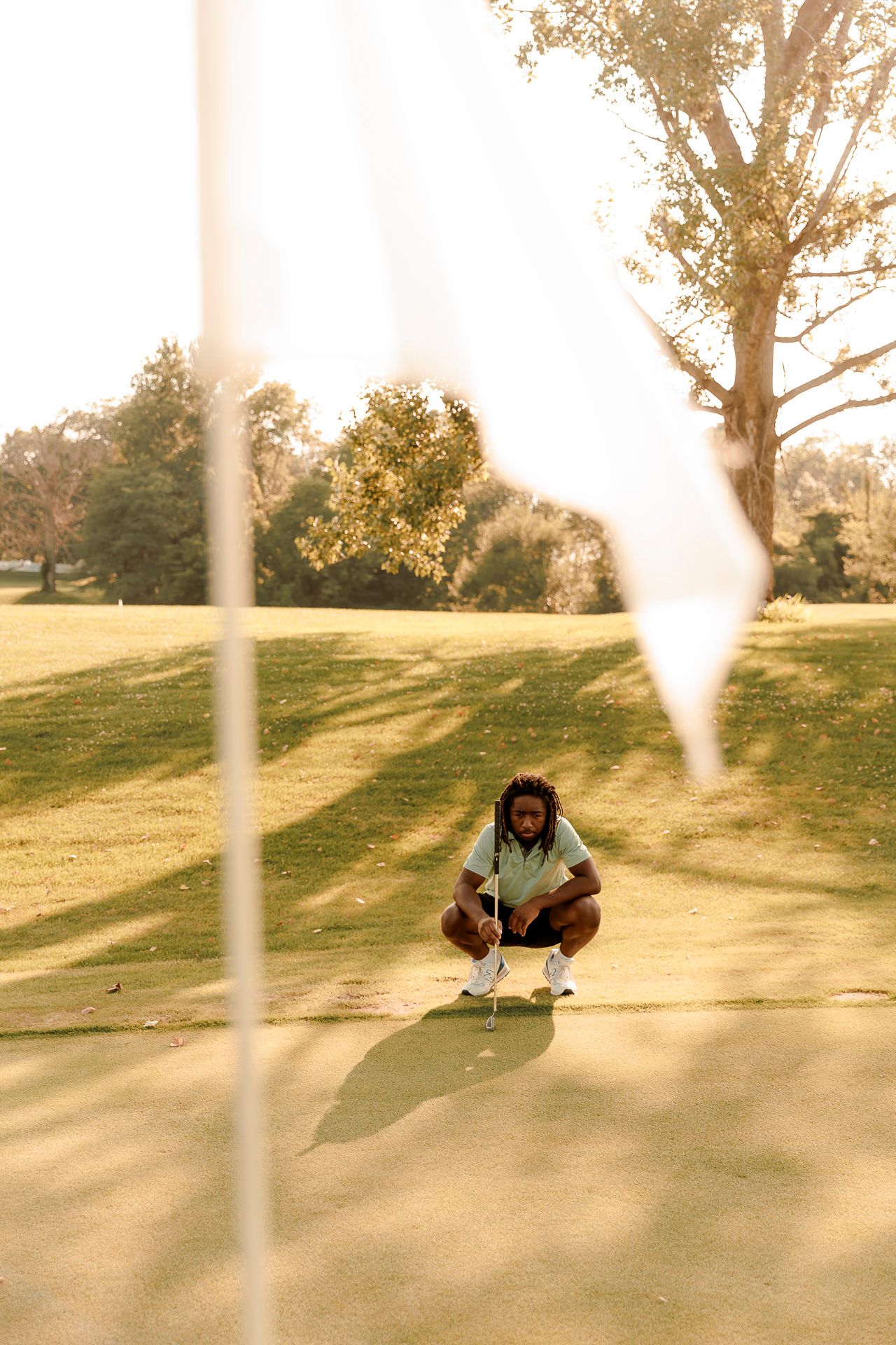 A golfer with dreadlocks crouches on a sunny golf course green, lining up a putt near a white flag.