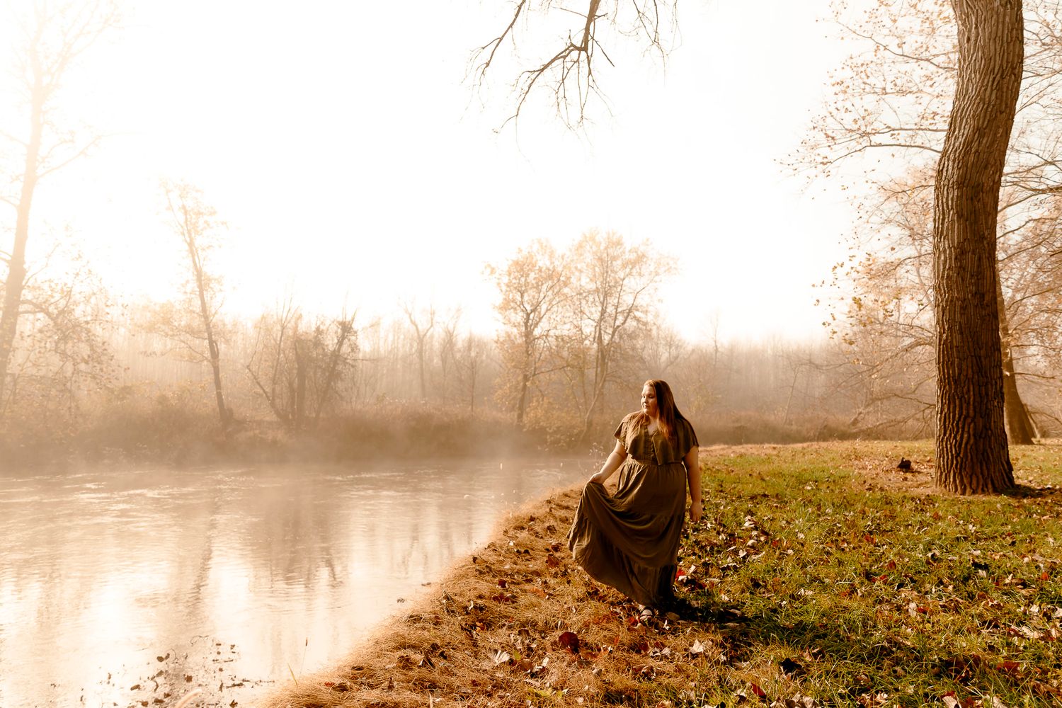 Person wrapped in blanket sitting by misty river in autumn park with bare trees and golden light.