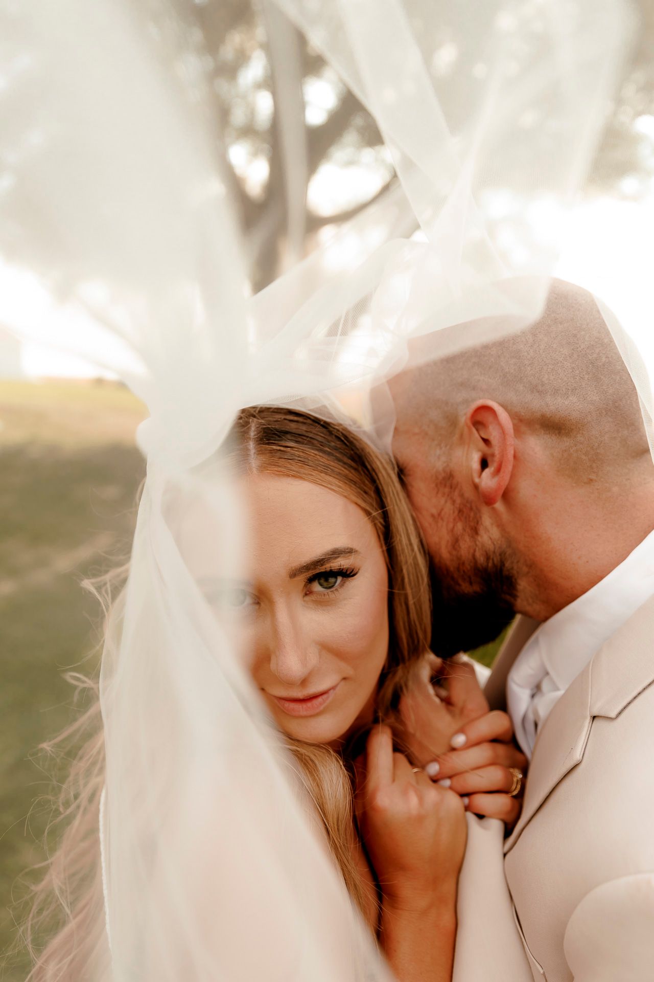 Bride and groom share intimate moment under flowing veil outdoors, bride gazing at camera with soft romantic expression.