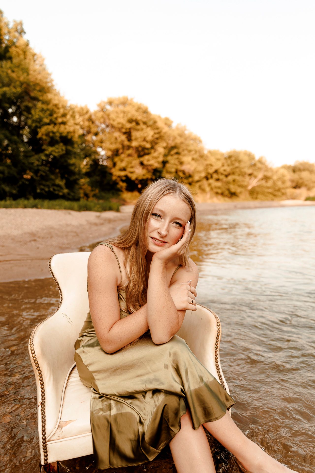 Young blonde woman in olive satin dress sitting in white chair by riverside with autumn trees in background.