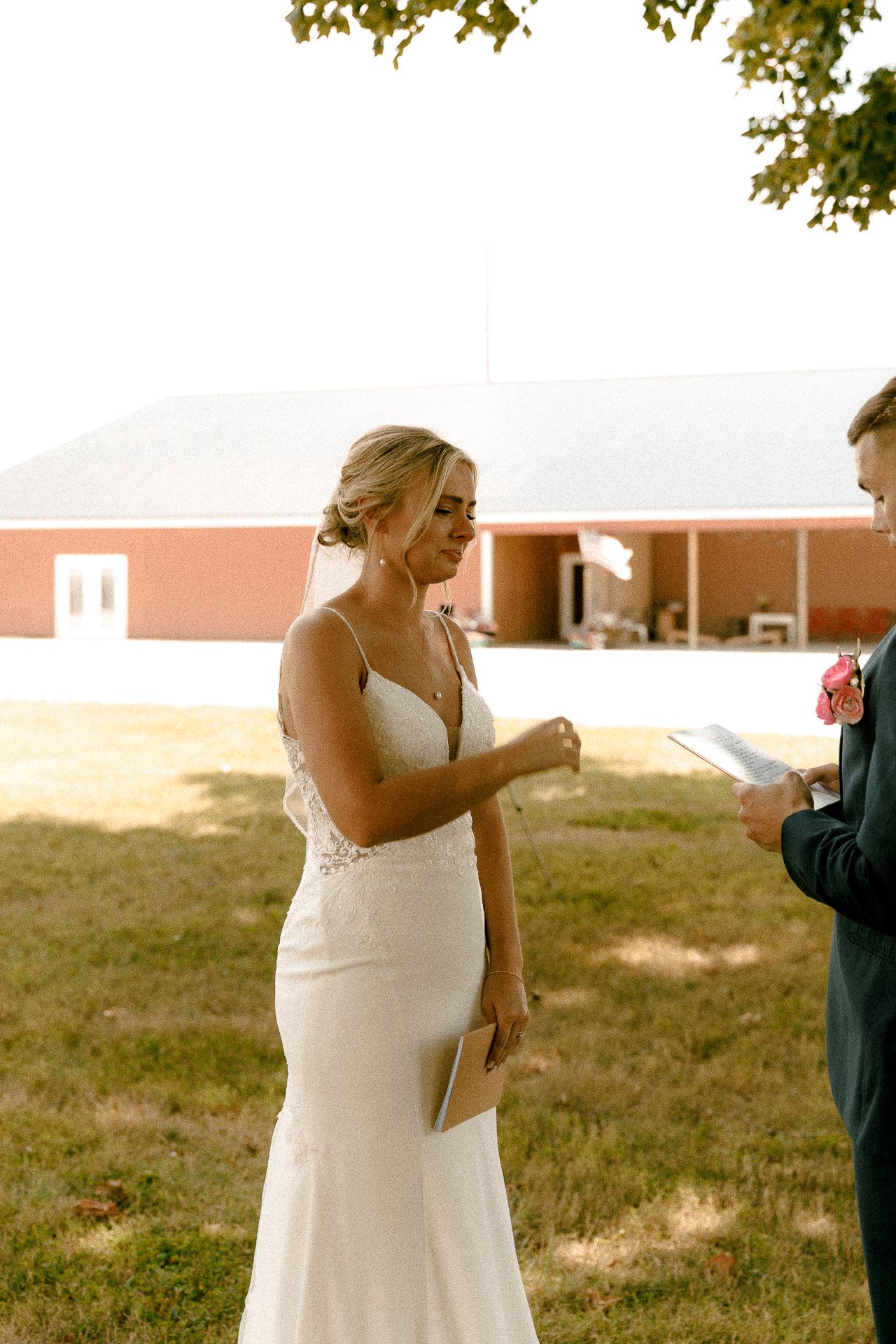 Bride in white lace gown reads vows outdoors near red barn during intimate wedding ceremony.