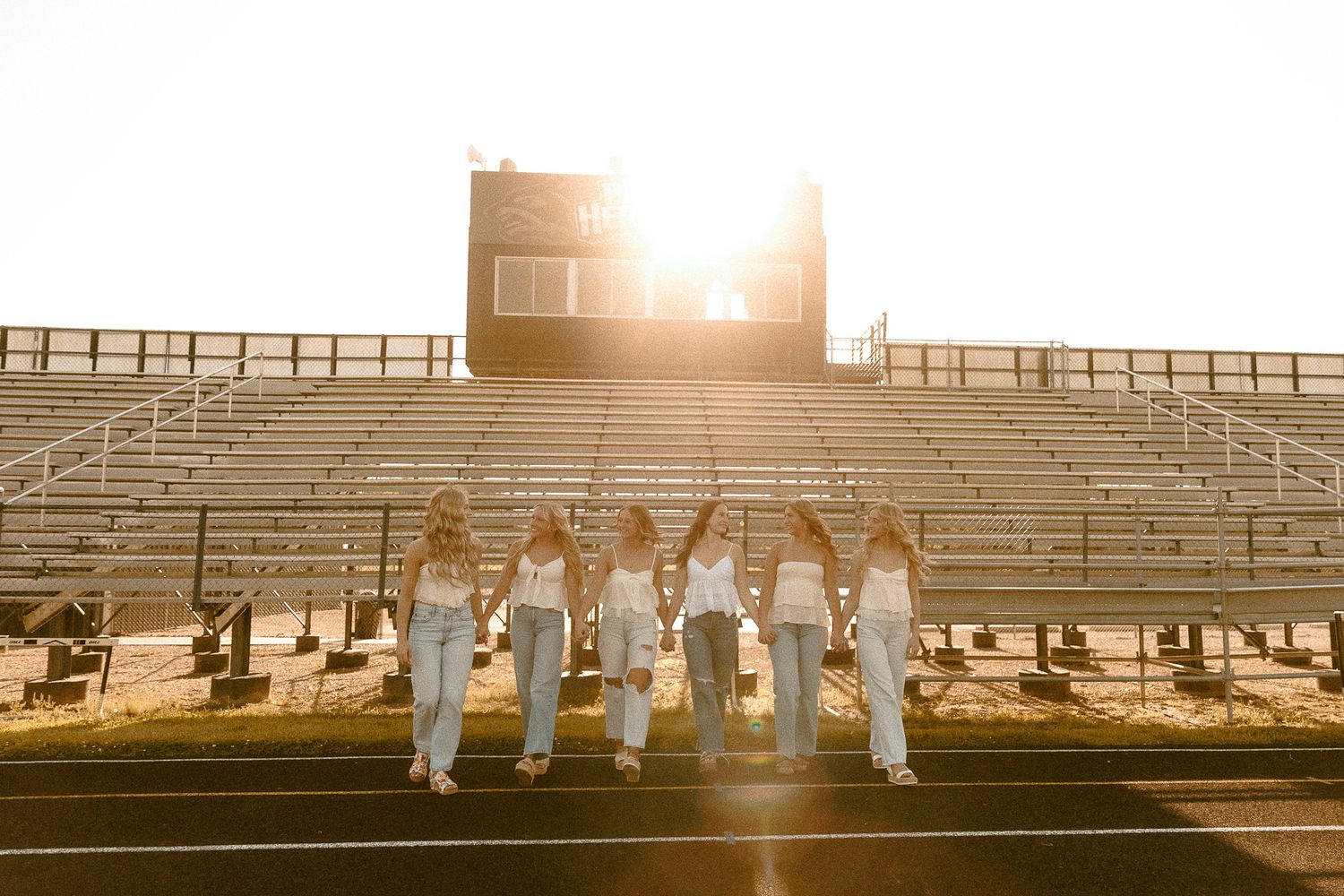 Seven girls in white tops and jeans walk together on a football field track at golden sunset hour.