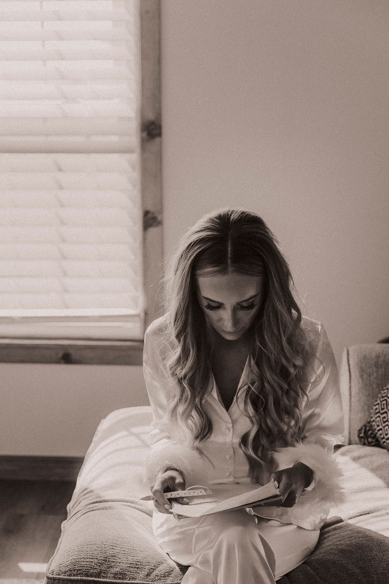 Black and white photo of a woman in white silk pajamas reading a book while sitting on a bed near a window.