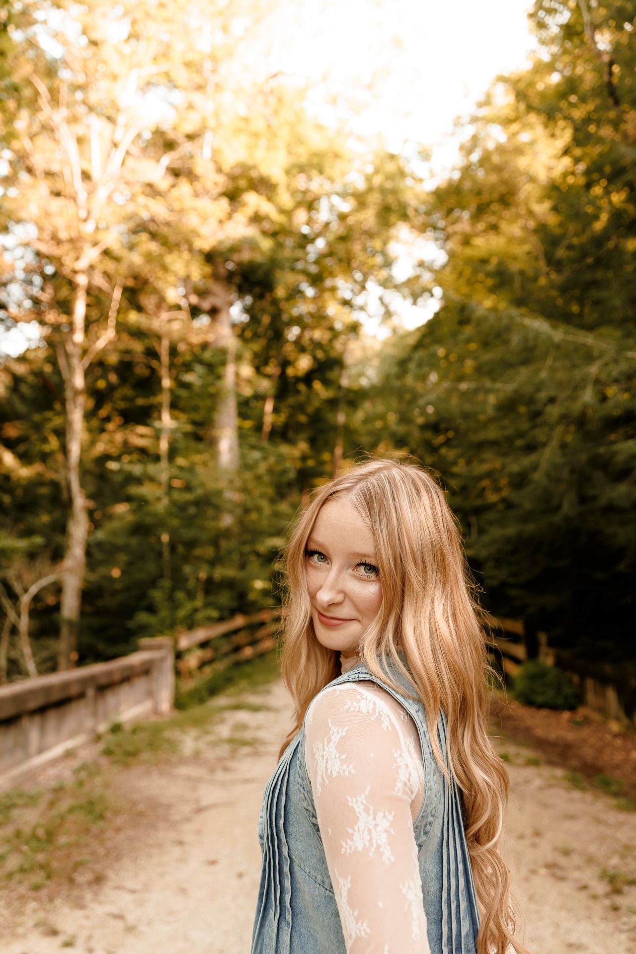 Young blonde woman smiling over shoulder on forest path in golden sunlight, wearing denim vest with lace detail.