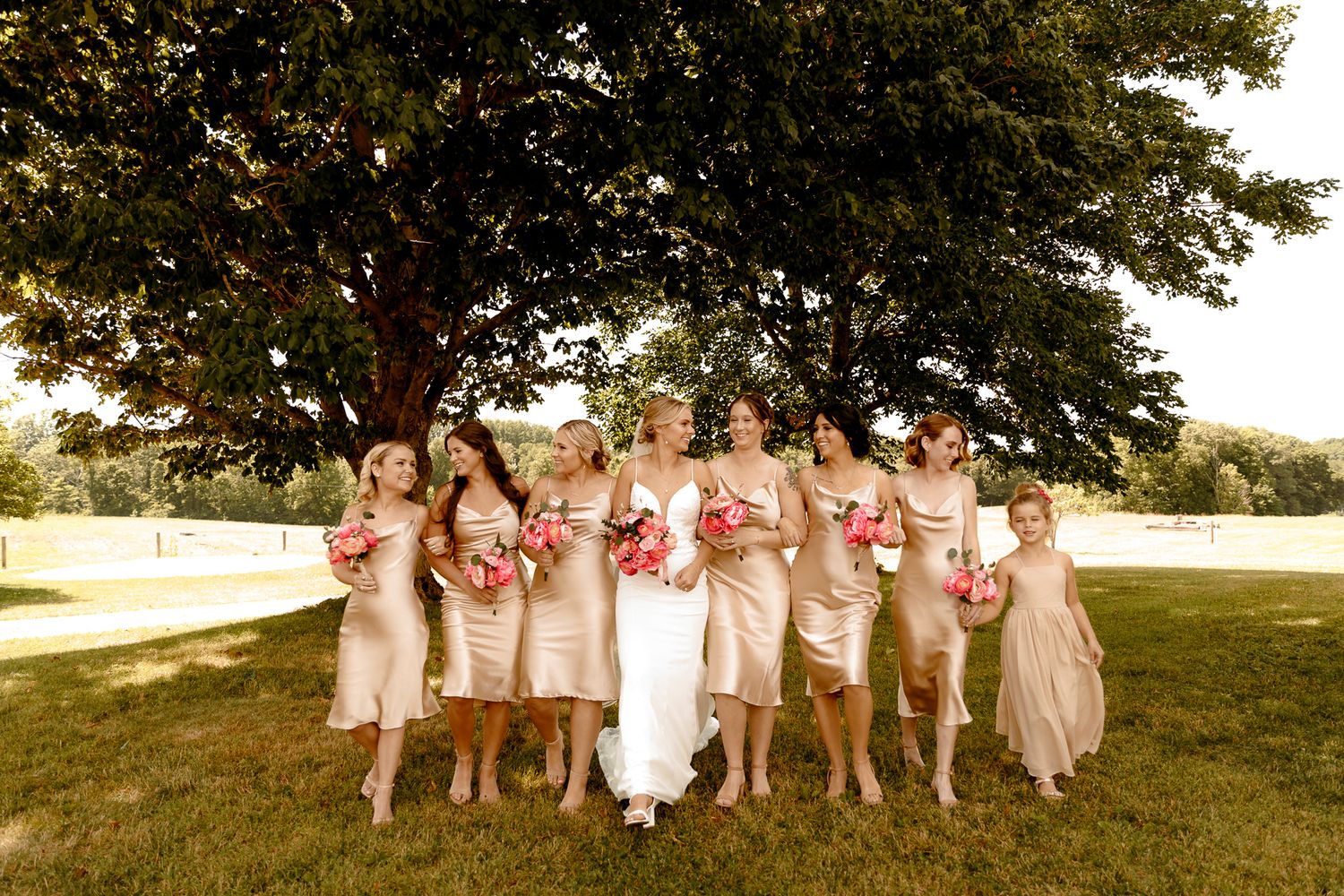 Bride in white gown walks with bridesmaids in champagne dresses holding pink bouquets under a large tree outdoors.