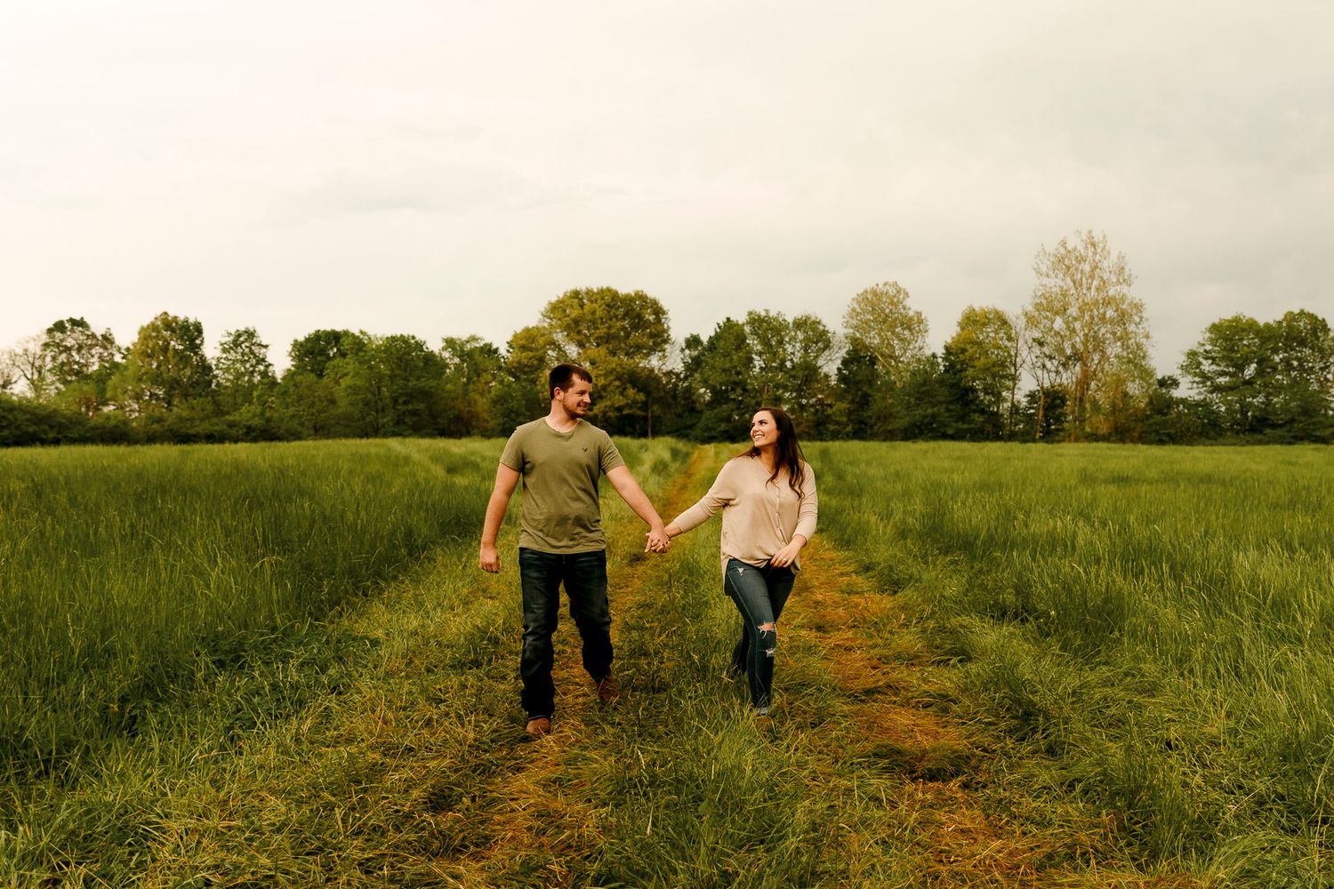 Couple holding hands walking through a lush green field on a cloudy day in a romantic outdoor setting.