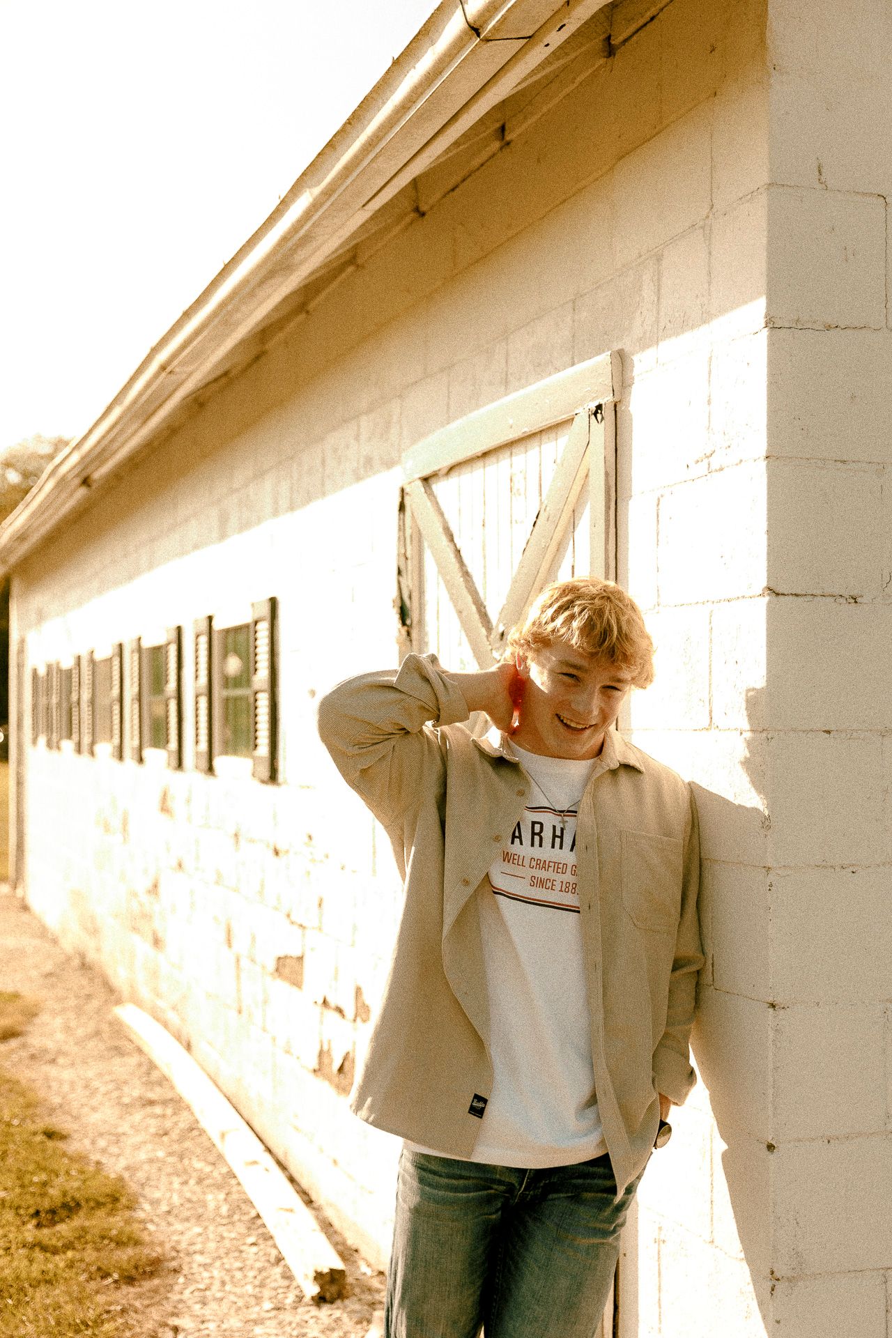 Young man in beige jacket and jeans smiling against a white brick barn wall in sunny outdoor setting.