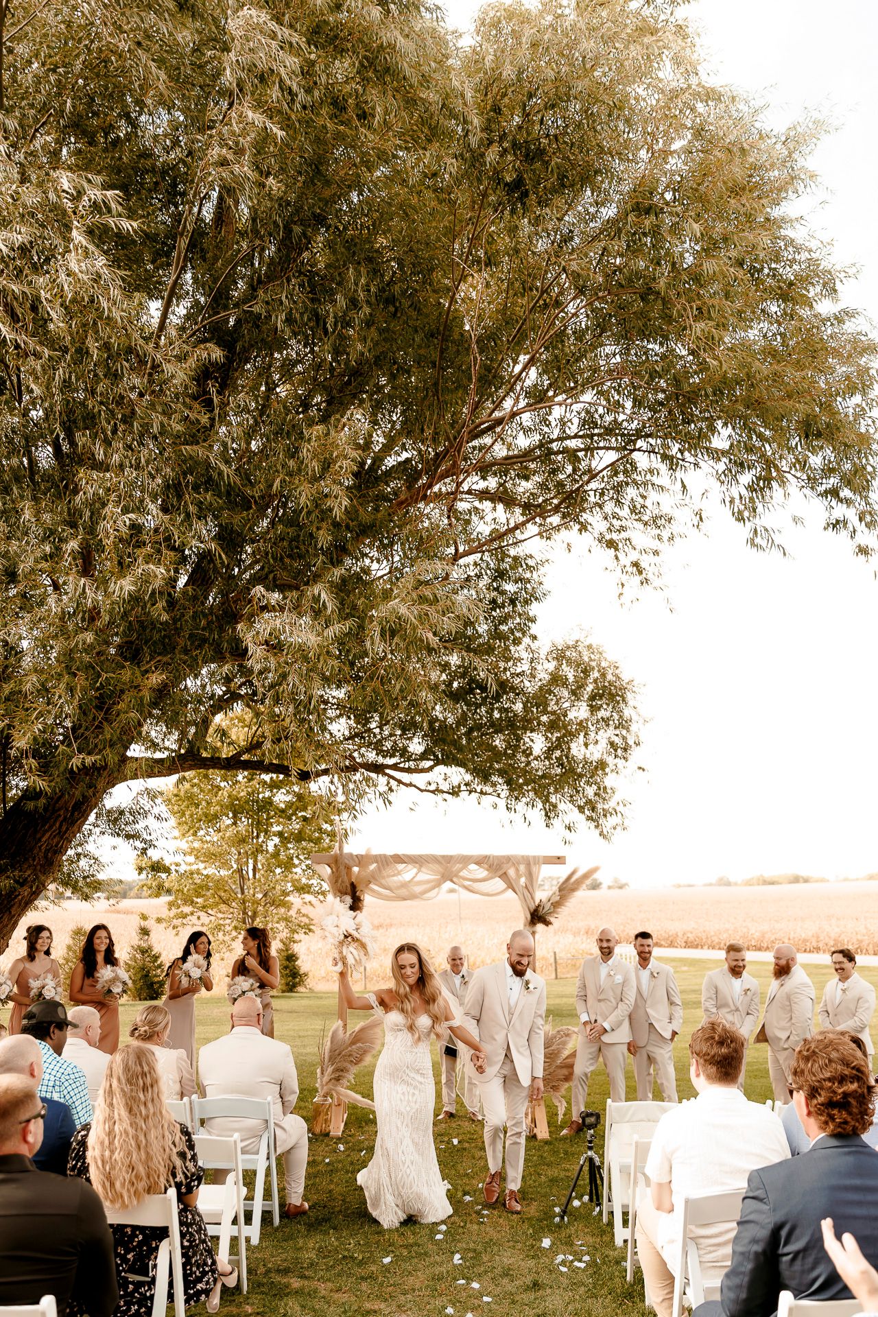 Outdoor wedding ceremony under a large tree with guests seated on white chairs as the couple walks down the aisle.
